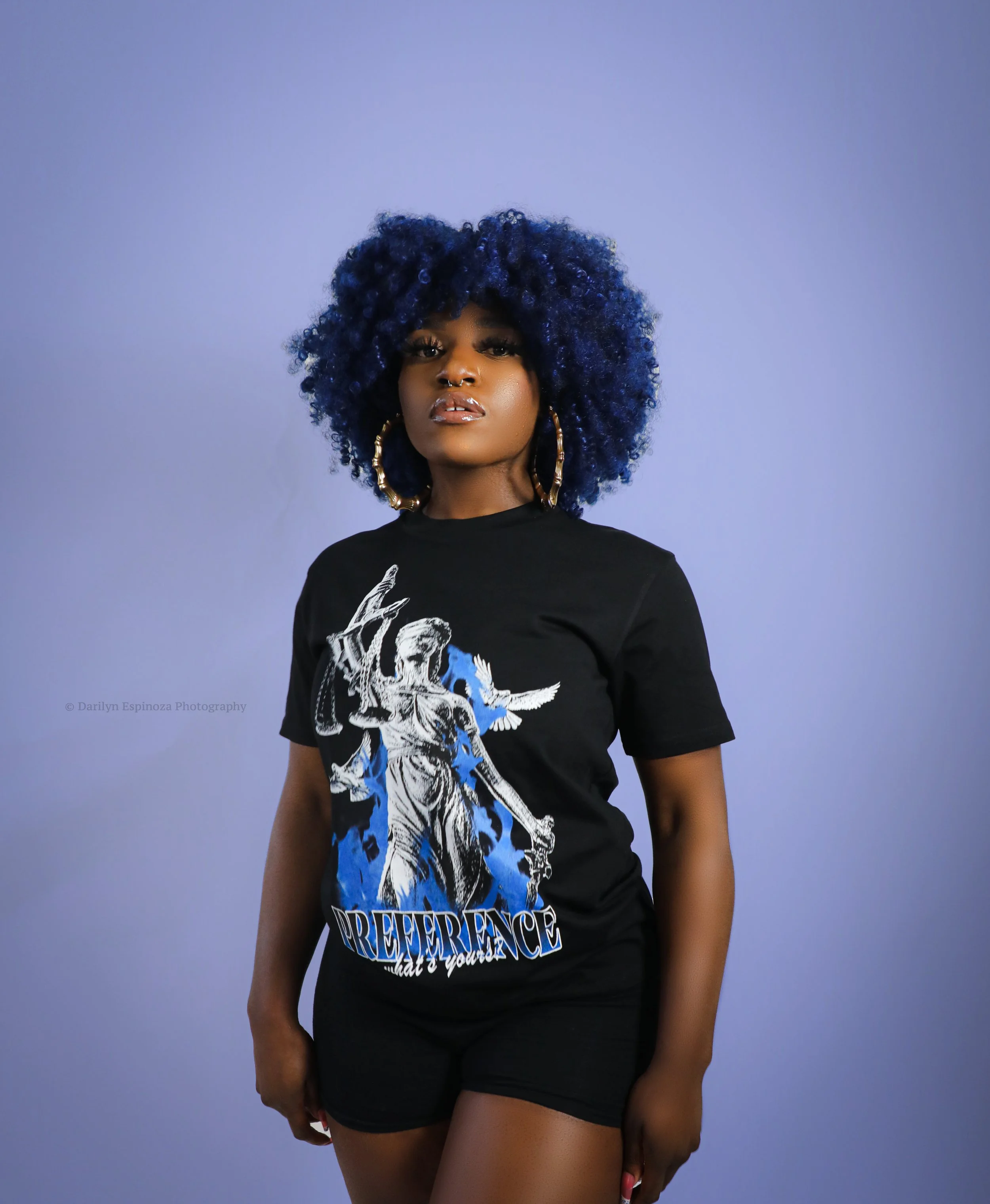 A woman with dark skin and large curly blue hair standing against a light purple background, wearing a black graphic T-shirt with an angel and dove design and the words 'PREFERENCE' on it, along with black shorts and large gold hoop earrings.