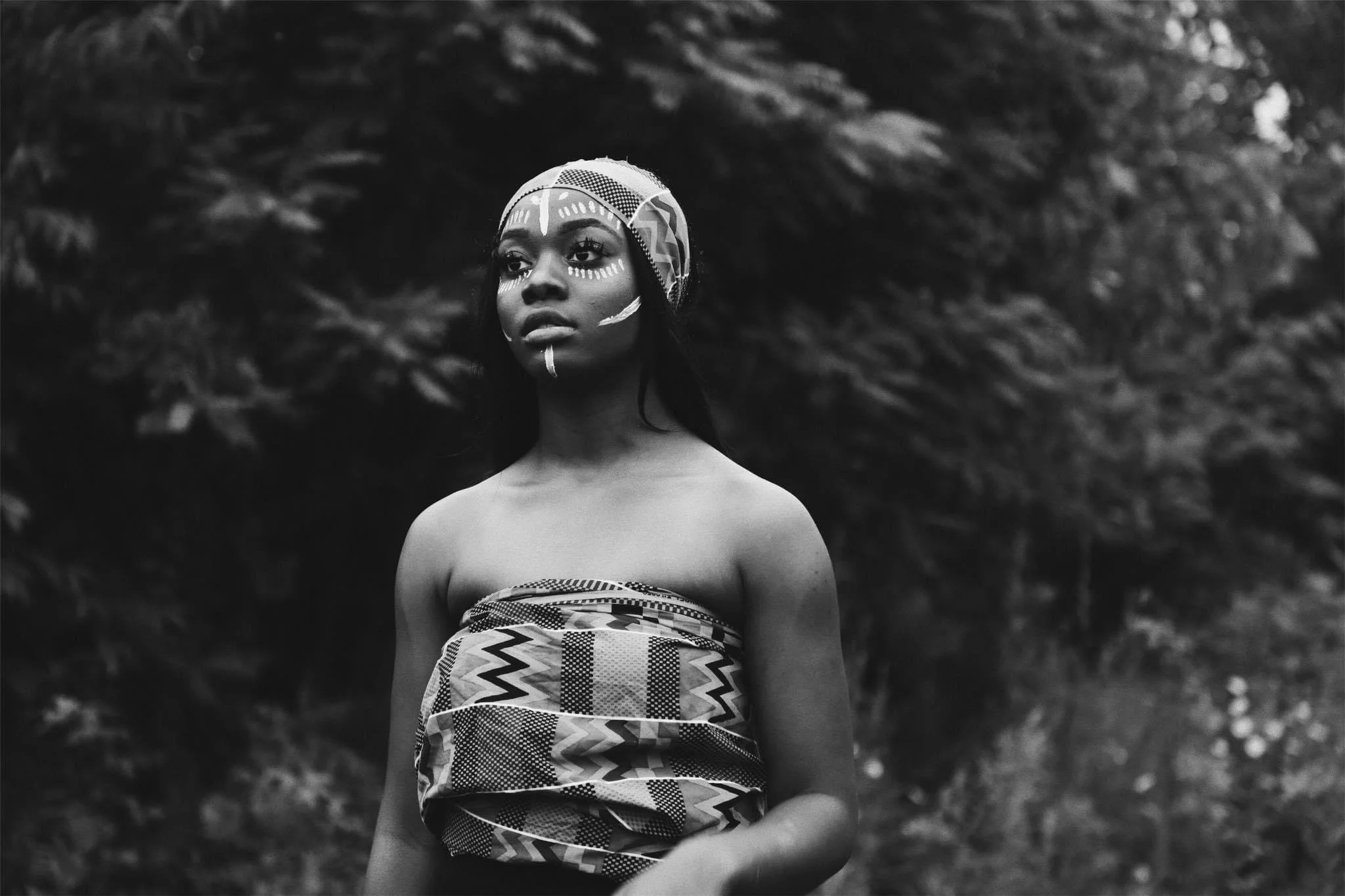 Black and white photo of woman wearing tribal face paint and head wrap, standing outdoors with dark foliage background.