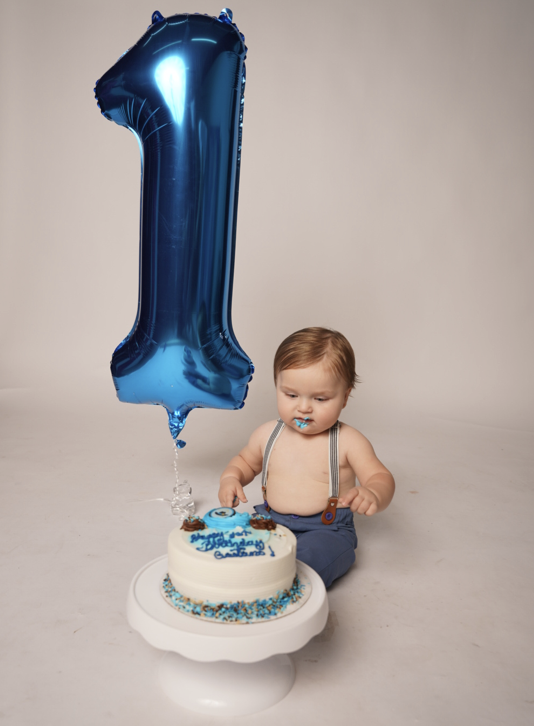 A young child sitting in front of a birthday cake with blue and white icing, a large blue balloon in the shape of the number one, and blue sprinkles, celebrating the child's first birthday.