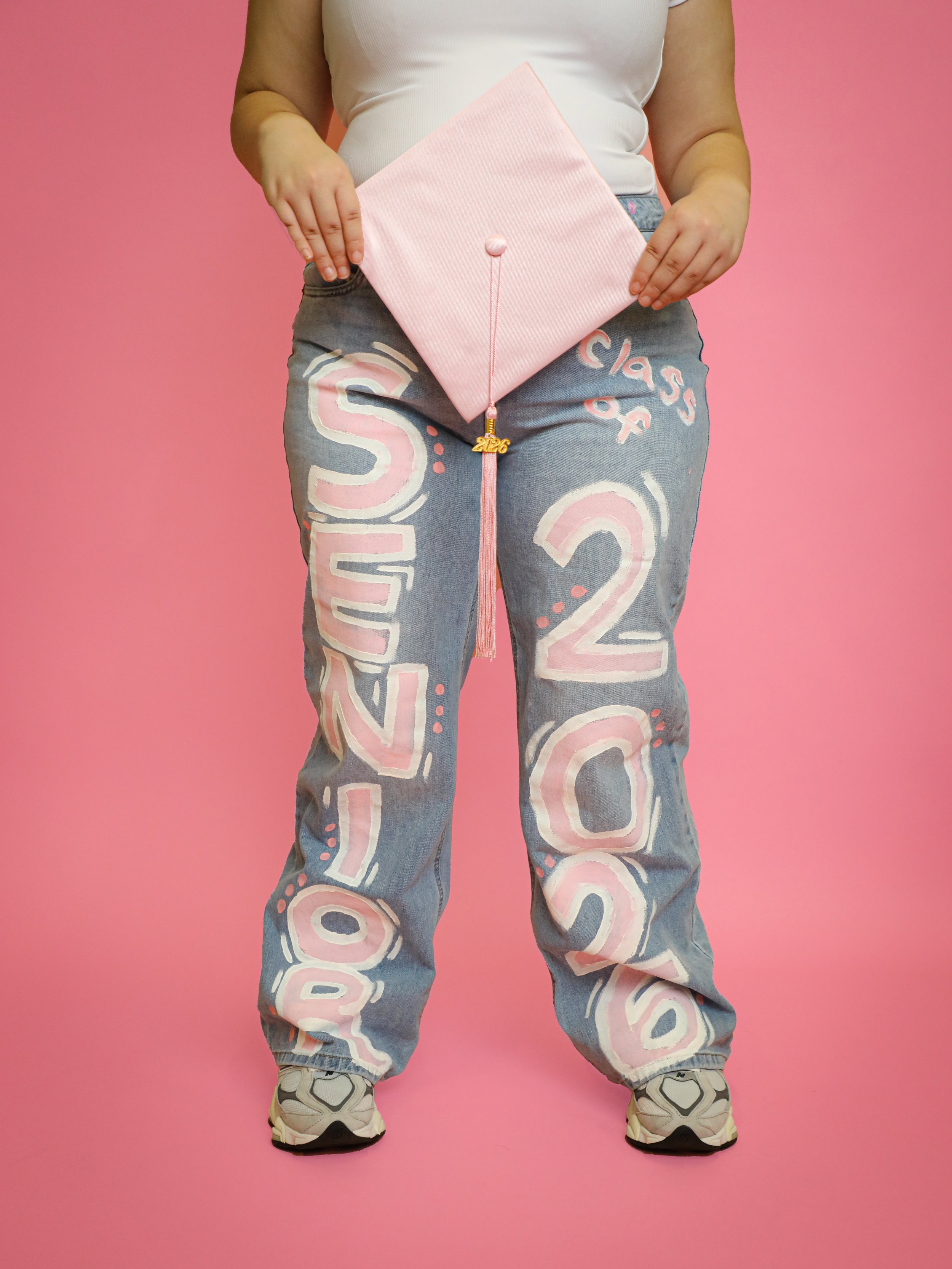 Person holding pink graduation cap with a tassel, standing against a pink background, wearing gray jeans with large pink and white lettering and sneakers.