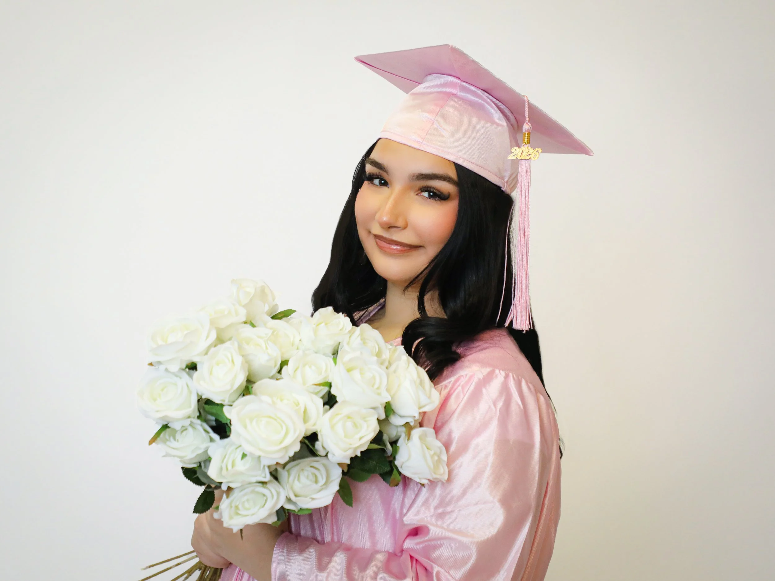 Graduating woman in pink cap and gown holding a bouquet of white roses, smiling.