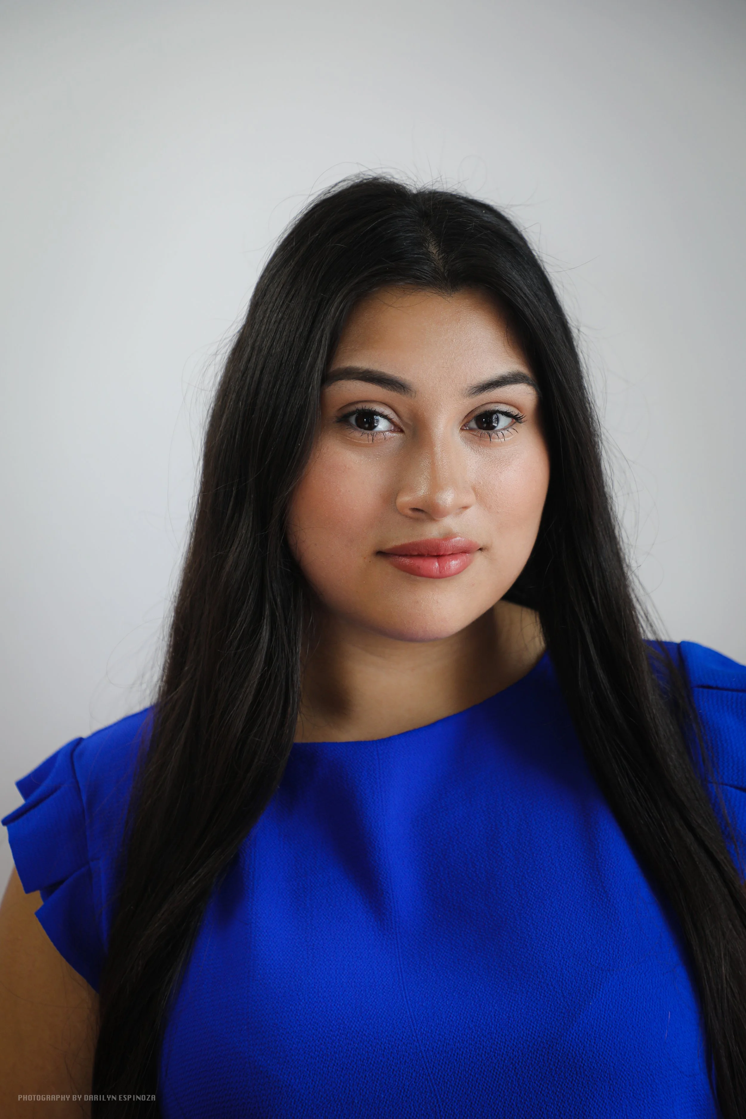 A young woman with long black hair, wearing a bright blue top, standing against a plain light gray background.