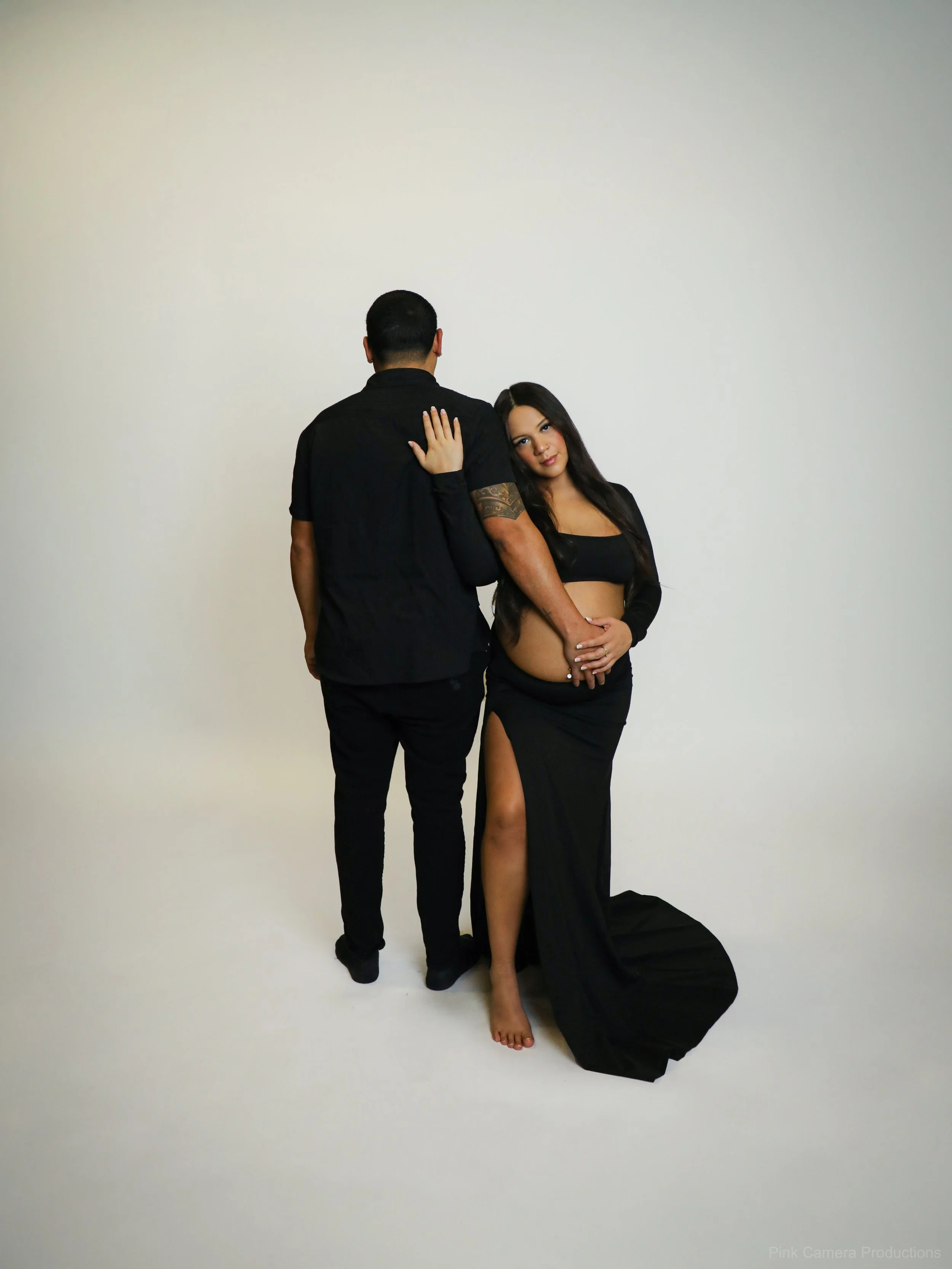A man and a woman pose together in front of a plain white background. The woman leans her head on the man's shoulder, and she is dressed in a black top and long black skirt with a high slit. The man is dressed in black, with his back to the camera.