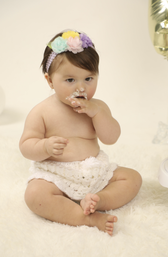 A baby girl sitting on a fluffy white rug, wearing a colorful flower headband and a white knitted diaper cover. She has food on her face and is eating with her fingers.