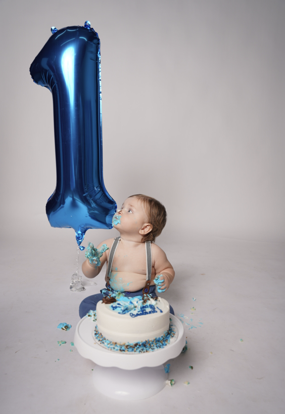 Baby sitting in front of a cake with blue and white icing, holding a blue balloon shaped as the number one, celebrating a first birthday.