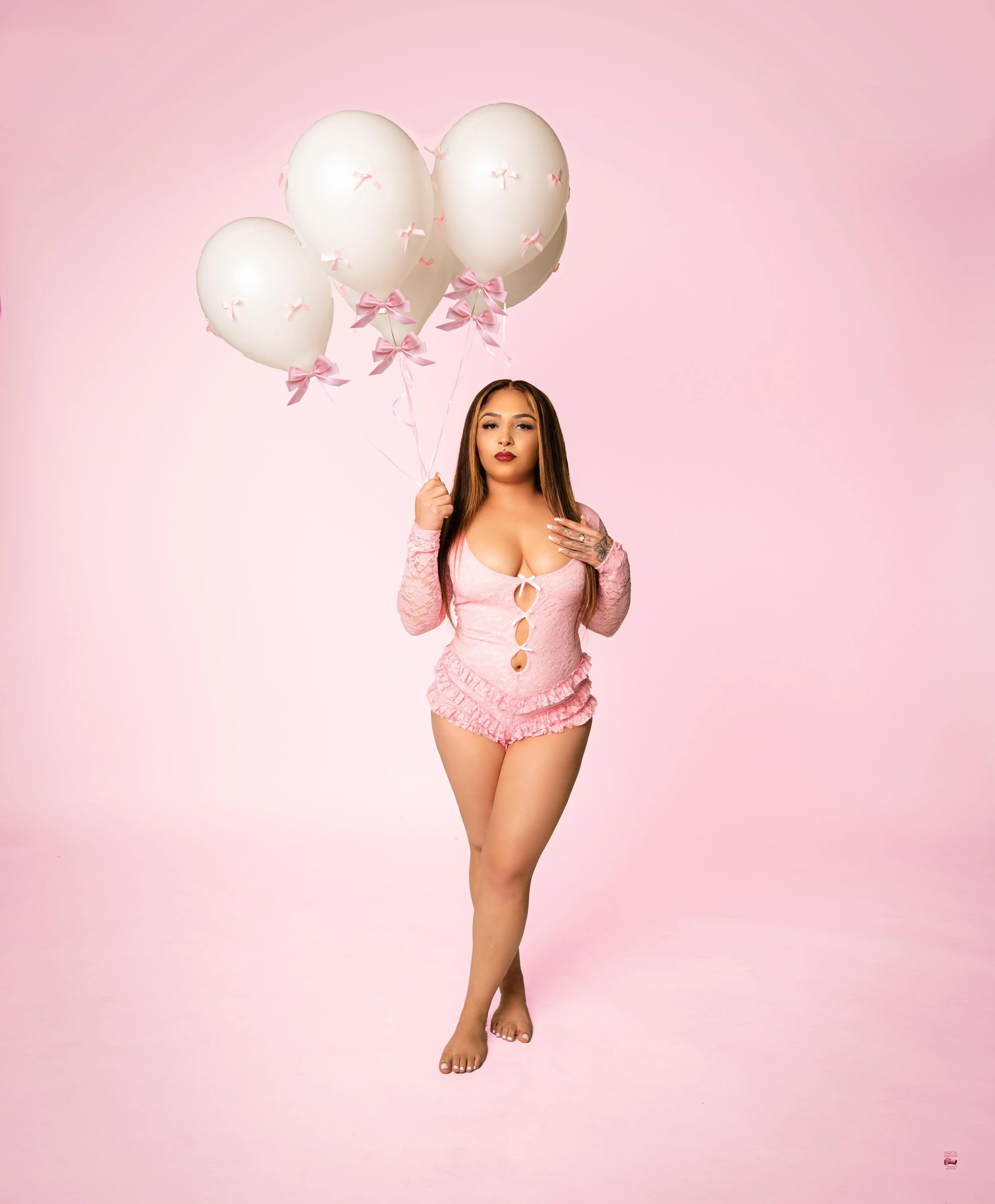 A woman standing barefoot against a pink background, wearing a pink ruffled two-piece outfit with cutouts, holding four white balloons decorated with small pink bows.