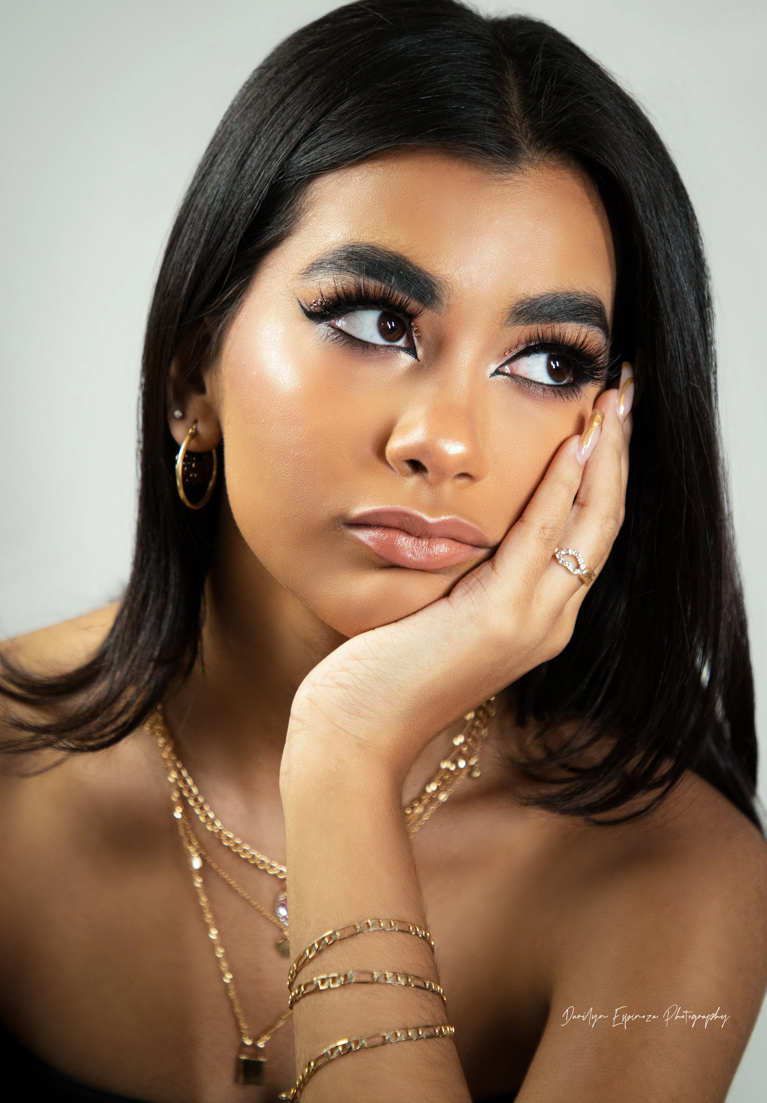 Close-up portrait of a woman with long black hair, tan skin, and gold jewelry, looking thoughtfully to the side with her hand resting on her face.