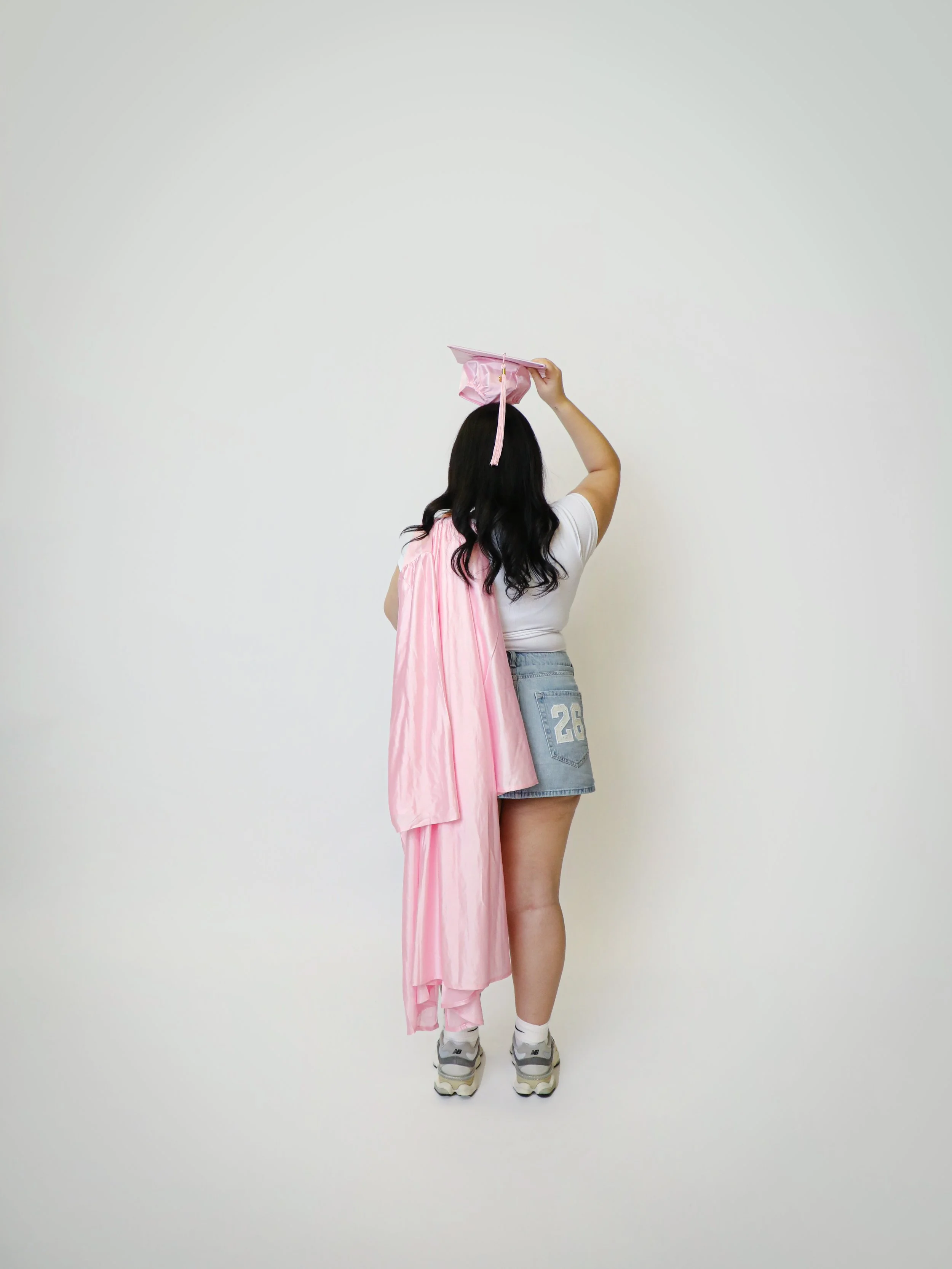 Person with long black hair, wearing a white t-shirt, denim shorts, and sneakers, holding a pink graduation cap and gown, standing against a plain white wall.