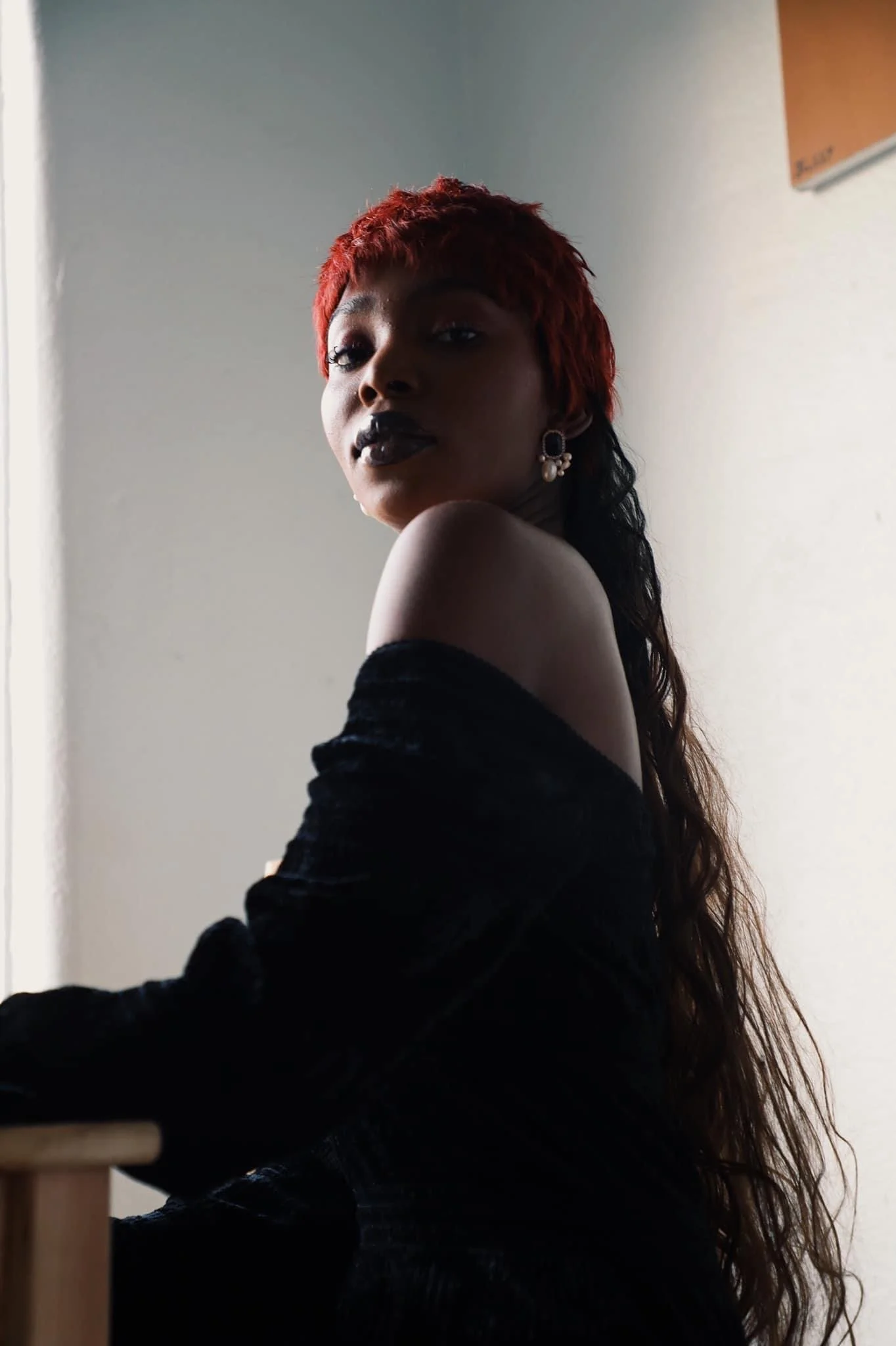 Portrait of a woman with red hair, black lipstick, wearing earrings and a black off-shoulder top, sitting indoors against a plain wall.