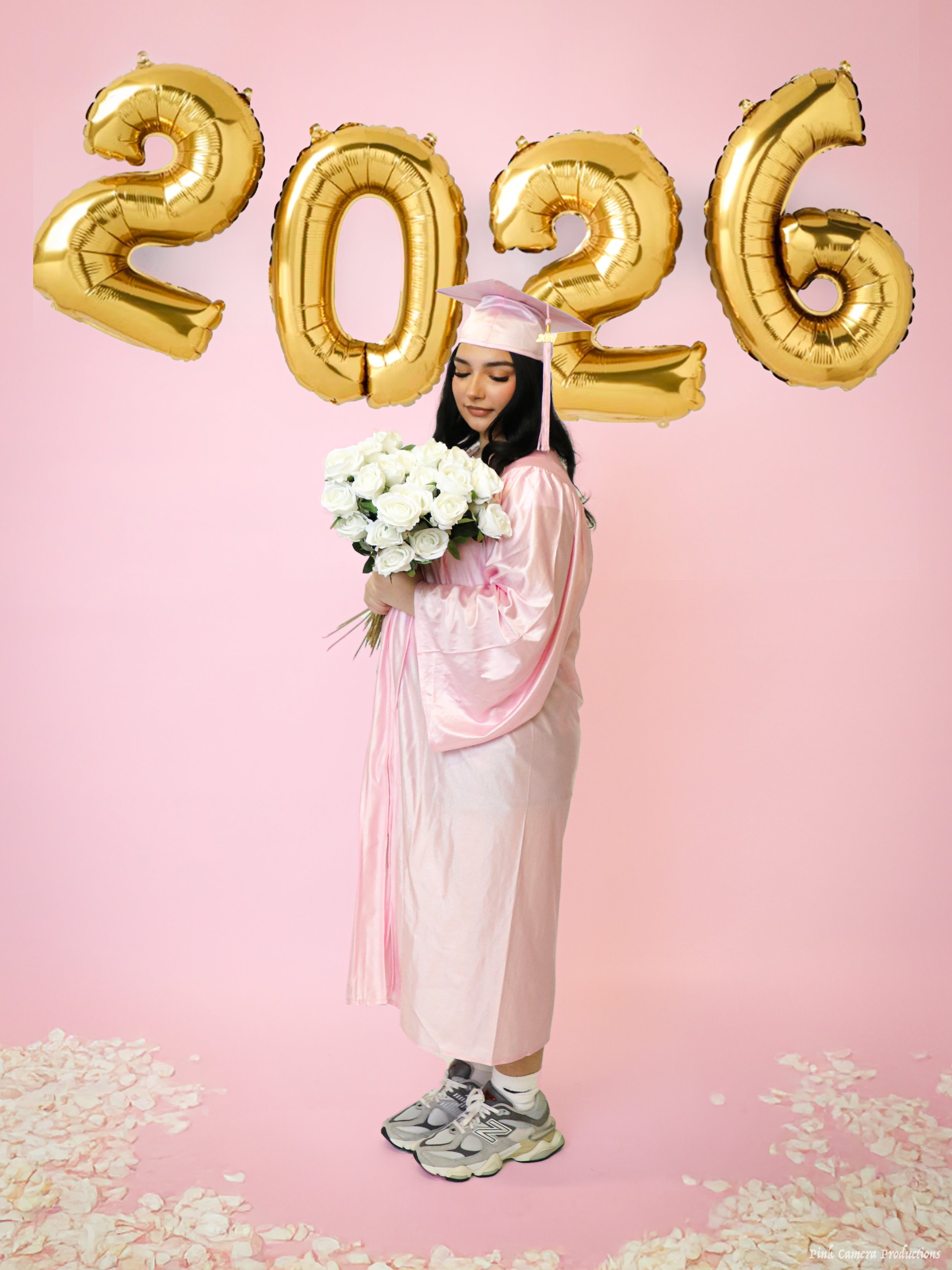 A young woman in a pink graduation gown and cap stands against a pink background, holding a bouquet of white roses. Gold balloons spelling out '2026' float behind her, and flower petals are scattered on the floor.