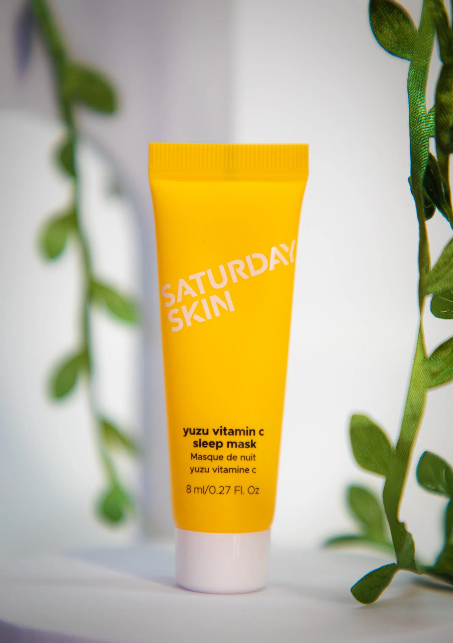 Yellow tube of SATURDAY SKIN yuzu vitamin C sleep mask on a white surface with green plant leaves on sides.