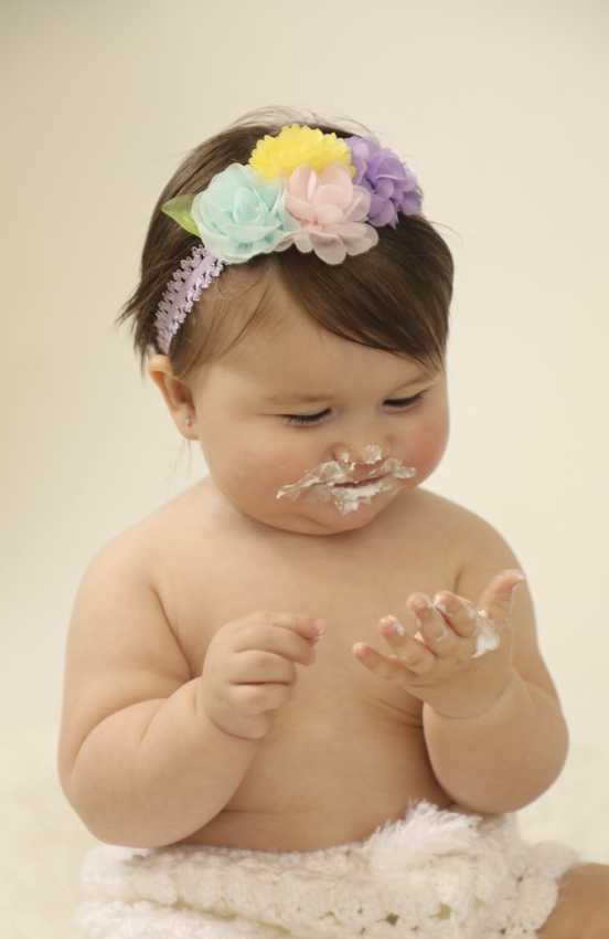 A young child with short brown hair wearing a colorful floral headband, looking at her hand with cake or cream on her face and hand, against a plain beige background.