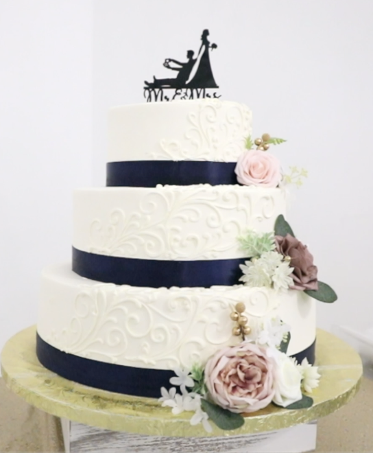 A four-tier wedding cake decorated with white swirling patterns, dark blue bands, and pink and white flowers, topped with a black silhouette cake topper of a bride and groom.