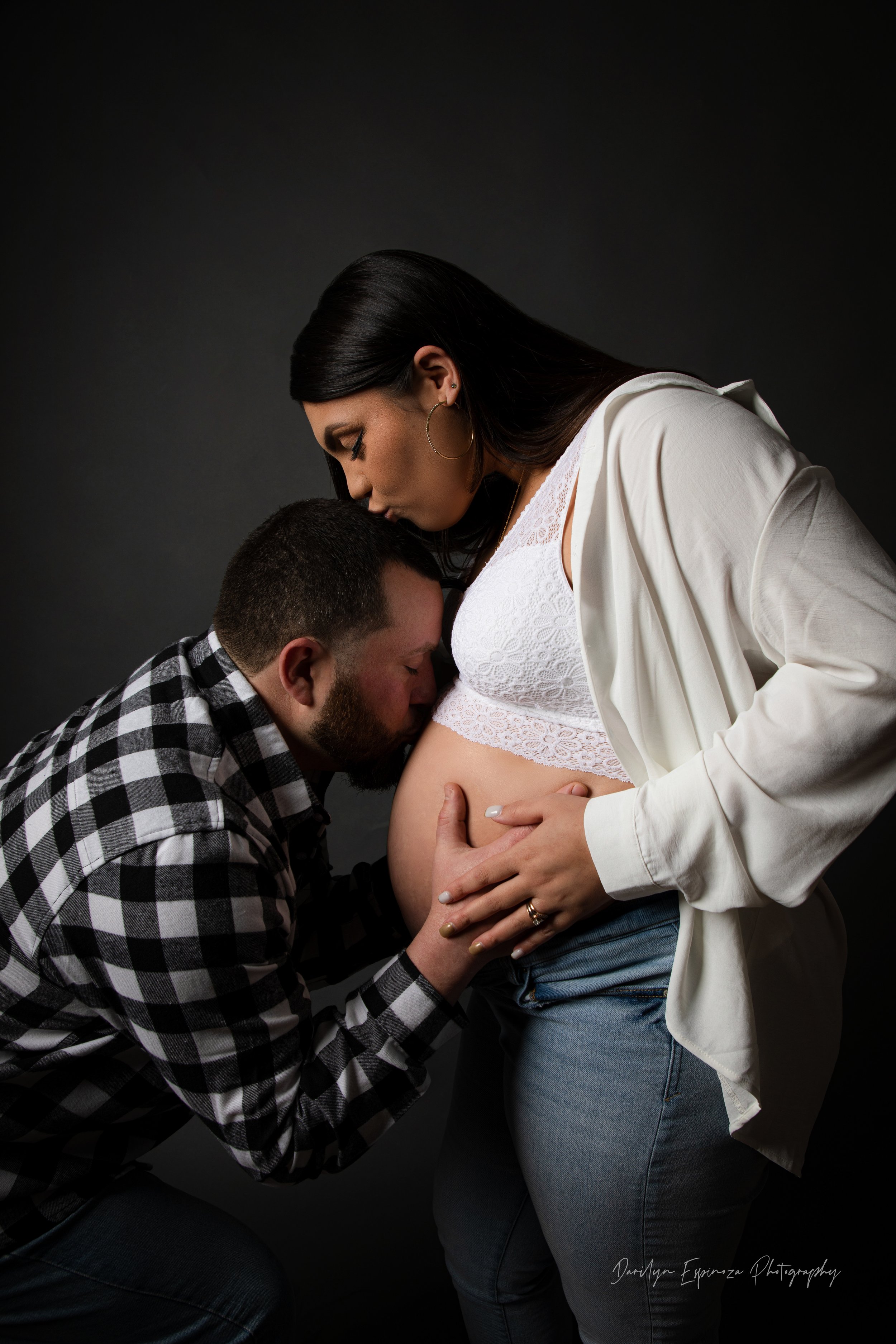 A pregnant woman with long dark hair, wearing a white lace top and light blue jeans, receiving a kiss on her belly from her partner, a man with short dark hair and a beard, dressed in a black and white checkered shirt. The woman is gently holding her