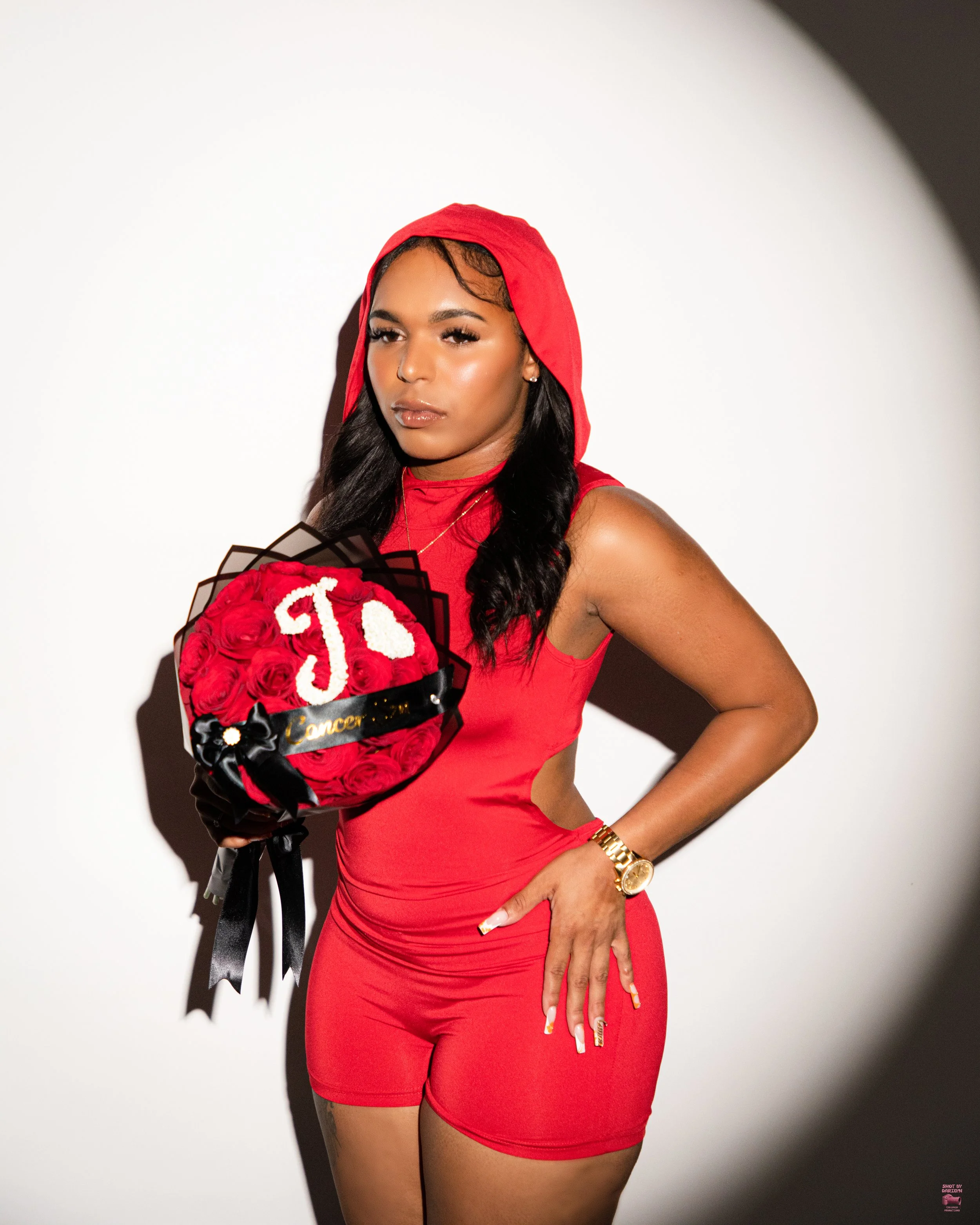 A woman in a red dress with a hood, holding a bouquet of red roses with a white letter 'J' on top, standing against a white background.