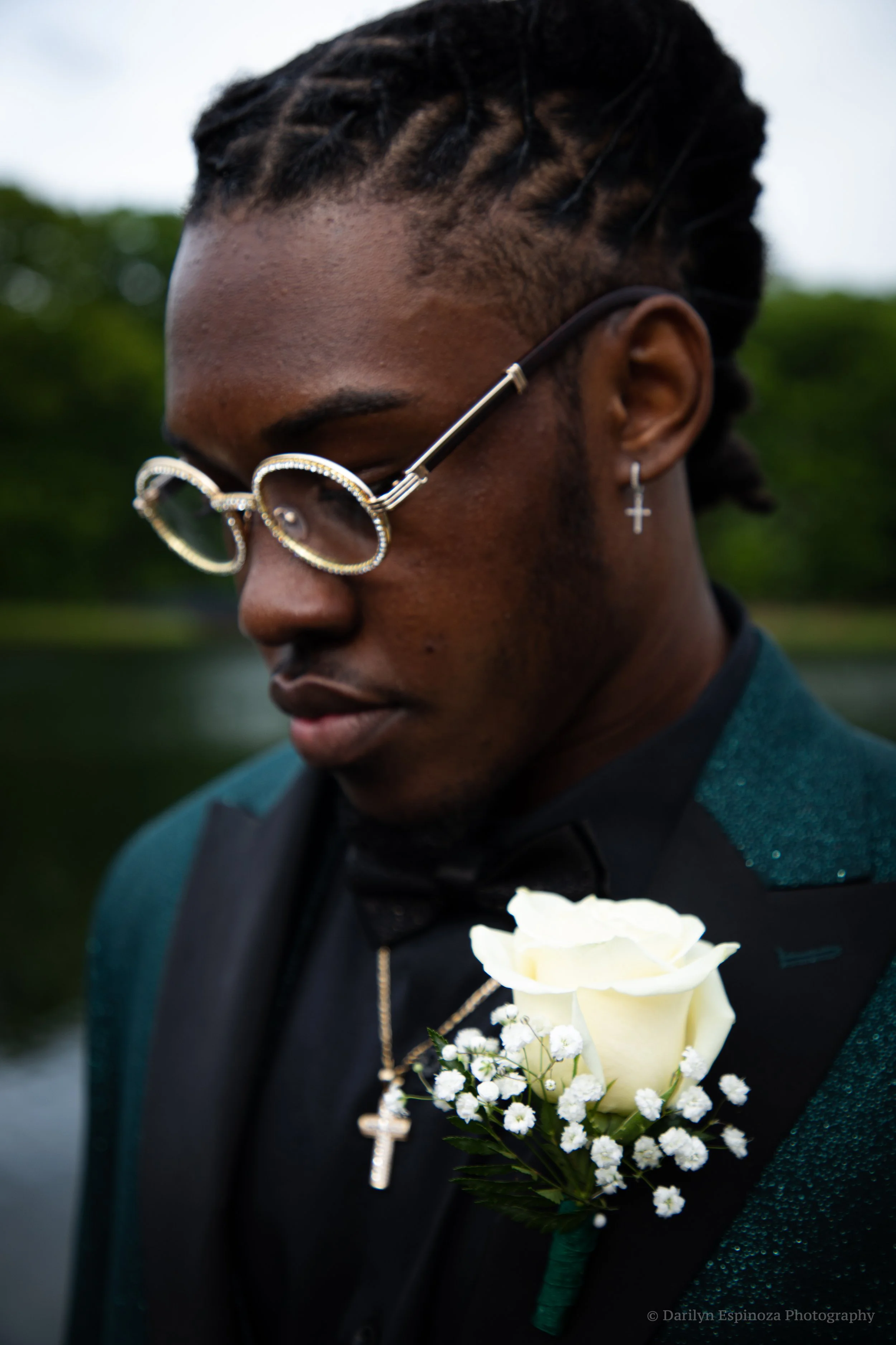 A young man with braided hair wearing glasses with rhinestones, a cross necklace, earrings, a suit with a boutonniere of white rose and baby's breath, and a dark shirt, looking down outdoors.