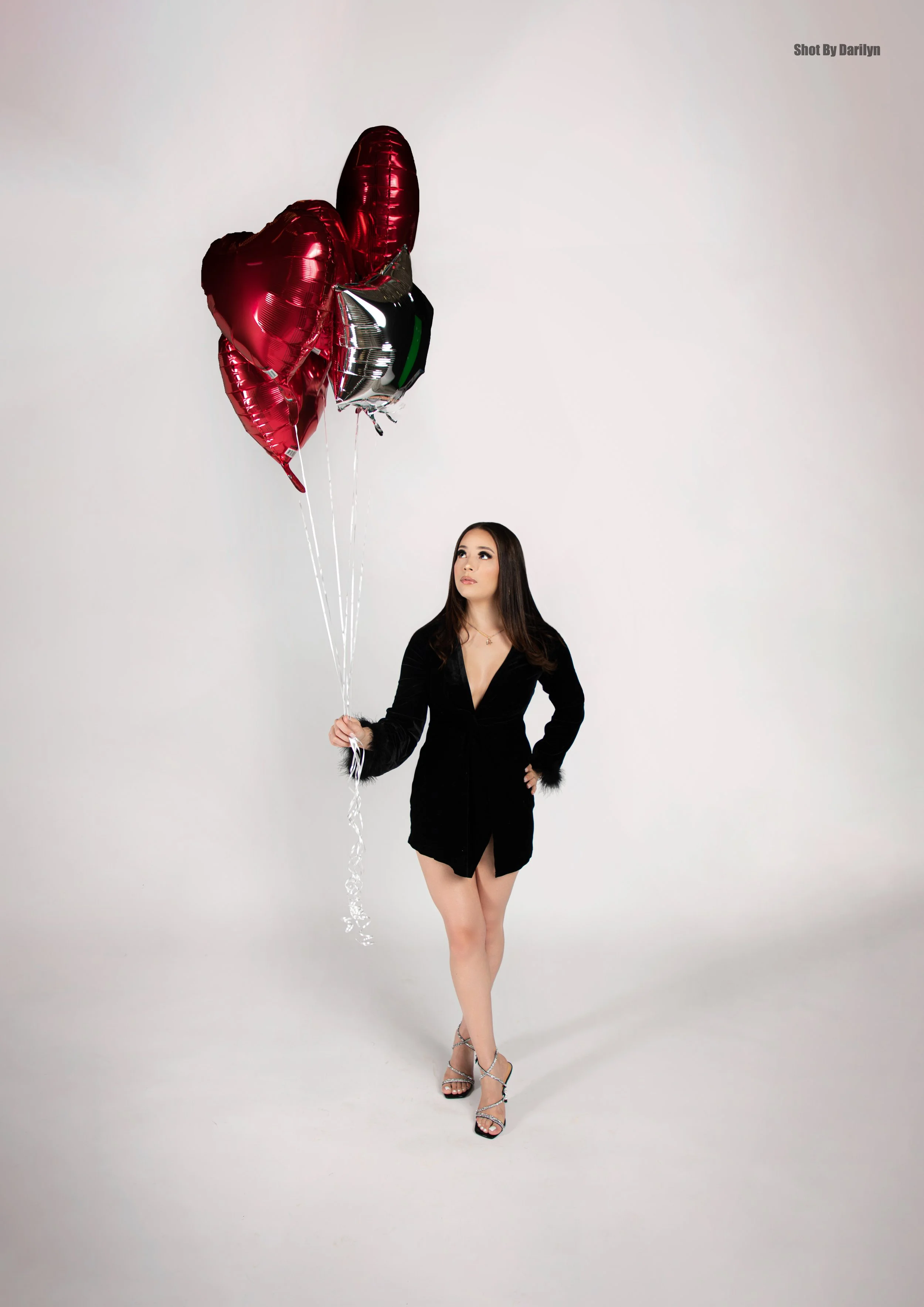 A woman in a black dress holding a bunch of red and silver heart-shaped balloons against a white background.