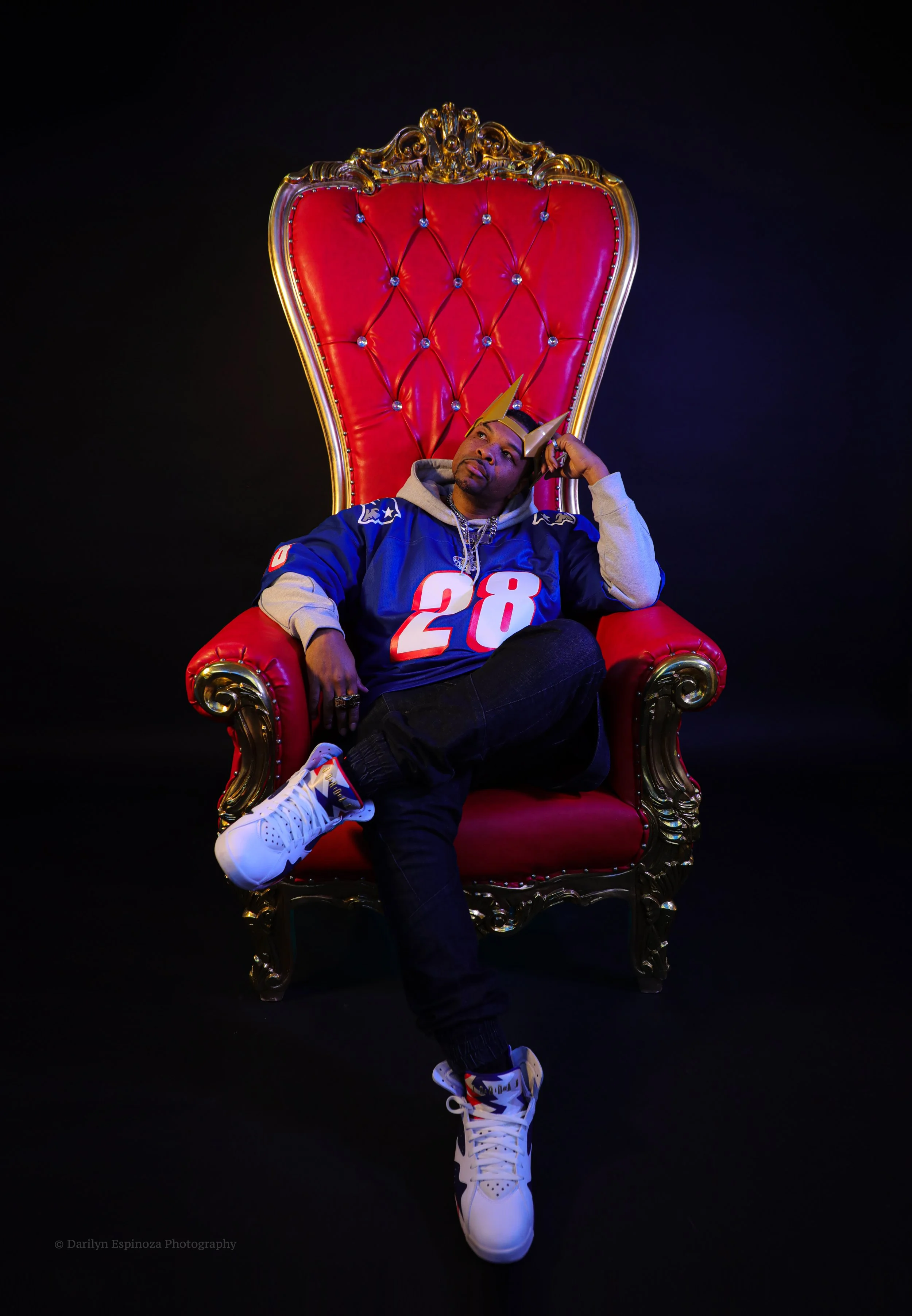 Man sitting on an ornate red and gold throne, wearing a blue sports jersey with the number 28, white sneakers, and unique horned headgear, against a black background.