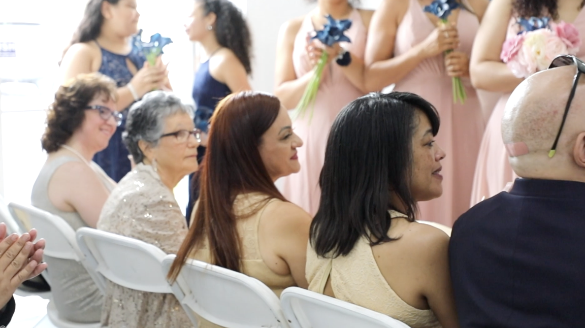 People attending a wedding or formal event, sitting and standing, holding flowers, dressed in elegant attire, in a bright indoor setting.