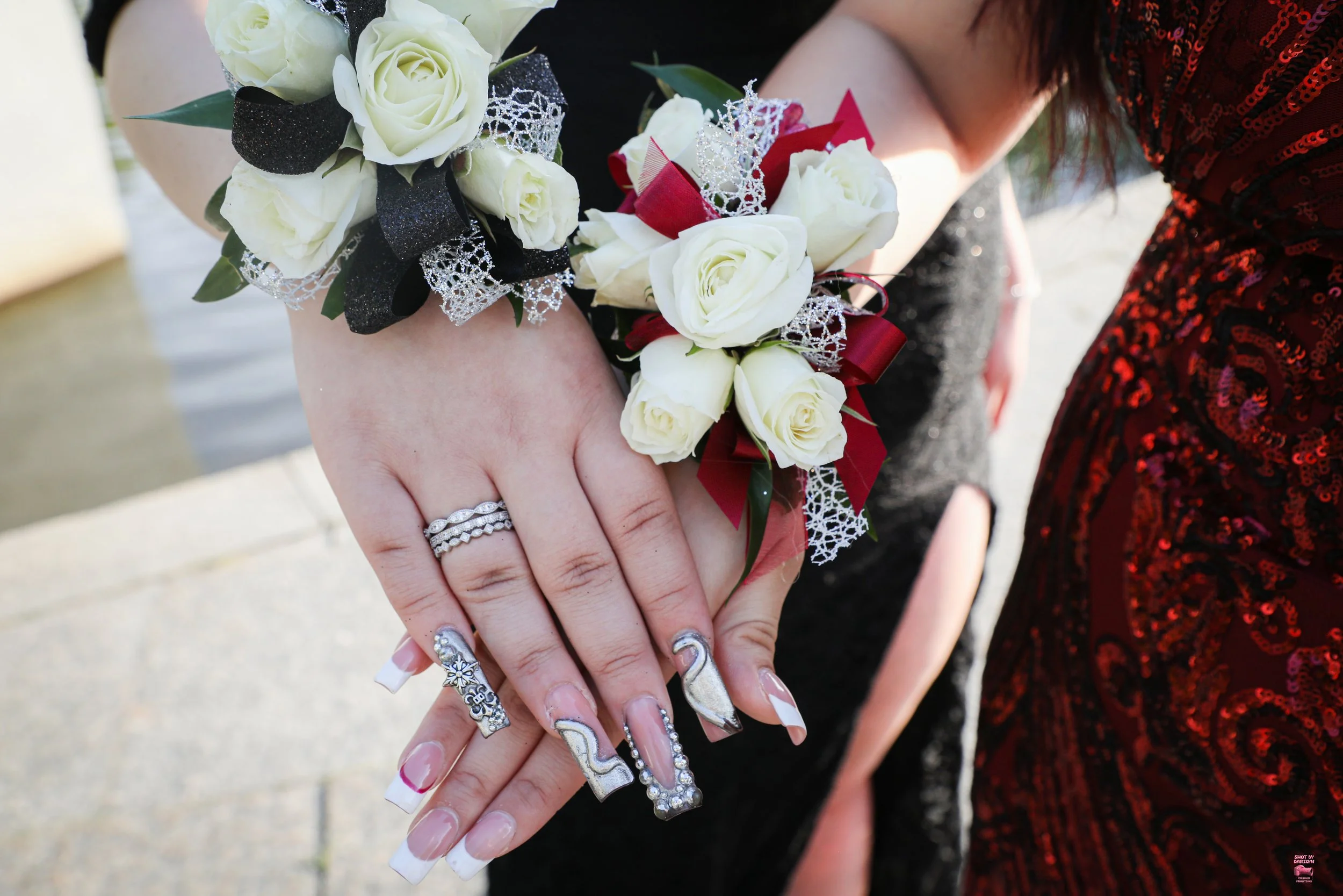 Close-up of a woman's hand with elaborate nail art and a wedding ring, holding a small bouquet of white roses with black and red ribbons and lace, with a person in a red dress in the background.