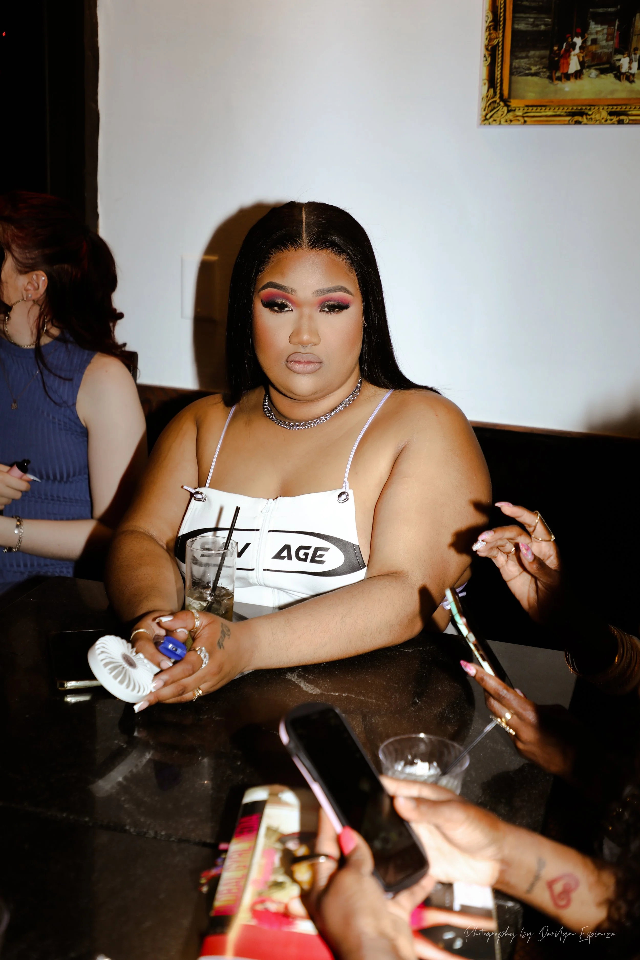 A woman with long black hair wearing a white dress with black letters, sitting at a table with her arms resting on it, holding a drink with a straw. She has makeup on, including red eyeshadow and neutral lipstick, and is wearing jewelry, including a 
