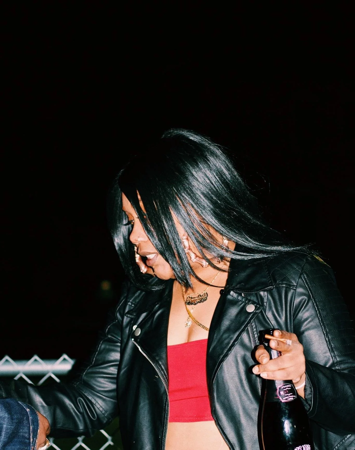 A woman with black, chin-length hair wearing a black leather jacket and red top, holding a dark bottle, looking down, at night.