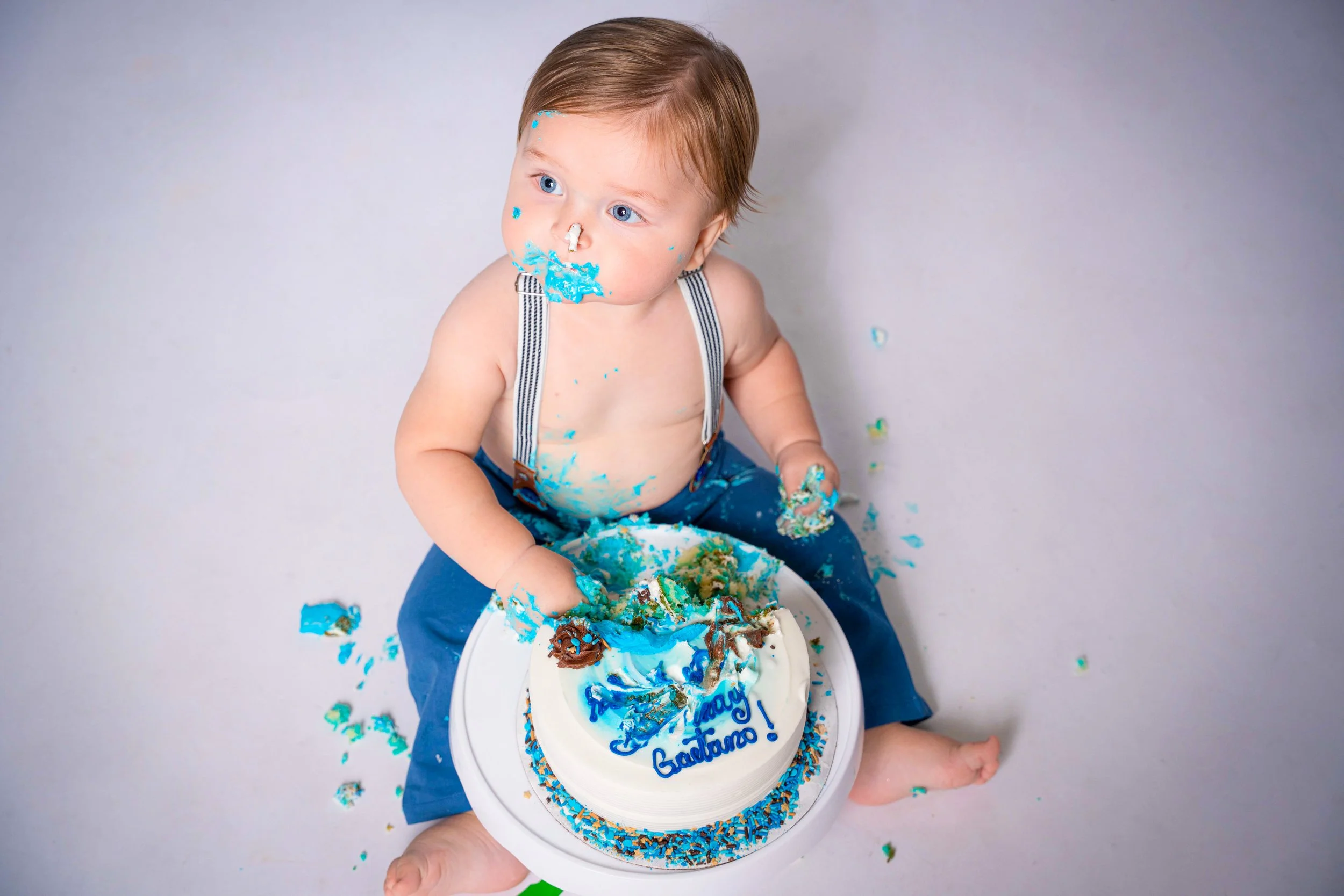 A young boy with blue eyes and brown hair sits on the floor with a blue and white birthday cake. The boy has blue cake and icing on his face and hands, and is wearing blue pants with suspenders, shirtless. The birthday cake has blue and white icing, 