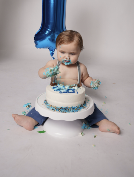 Baby sitting on a white potty chair with a cake on top, covered in blue and white icing, and a blue balloon shaped like the number one in the background, celebrating a first birthday.