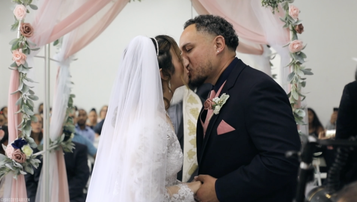 A bride and groom kiss during a wedding ceremony, standing under a pink and white floral arch with guests in the background.