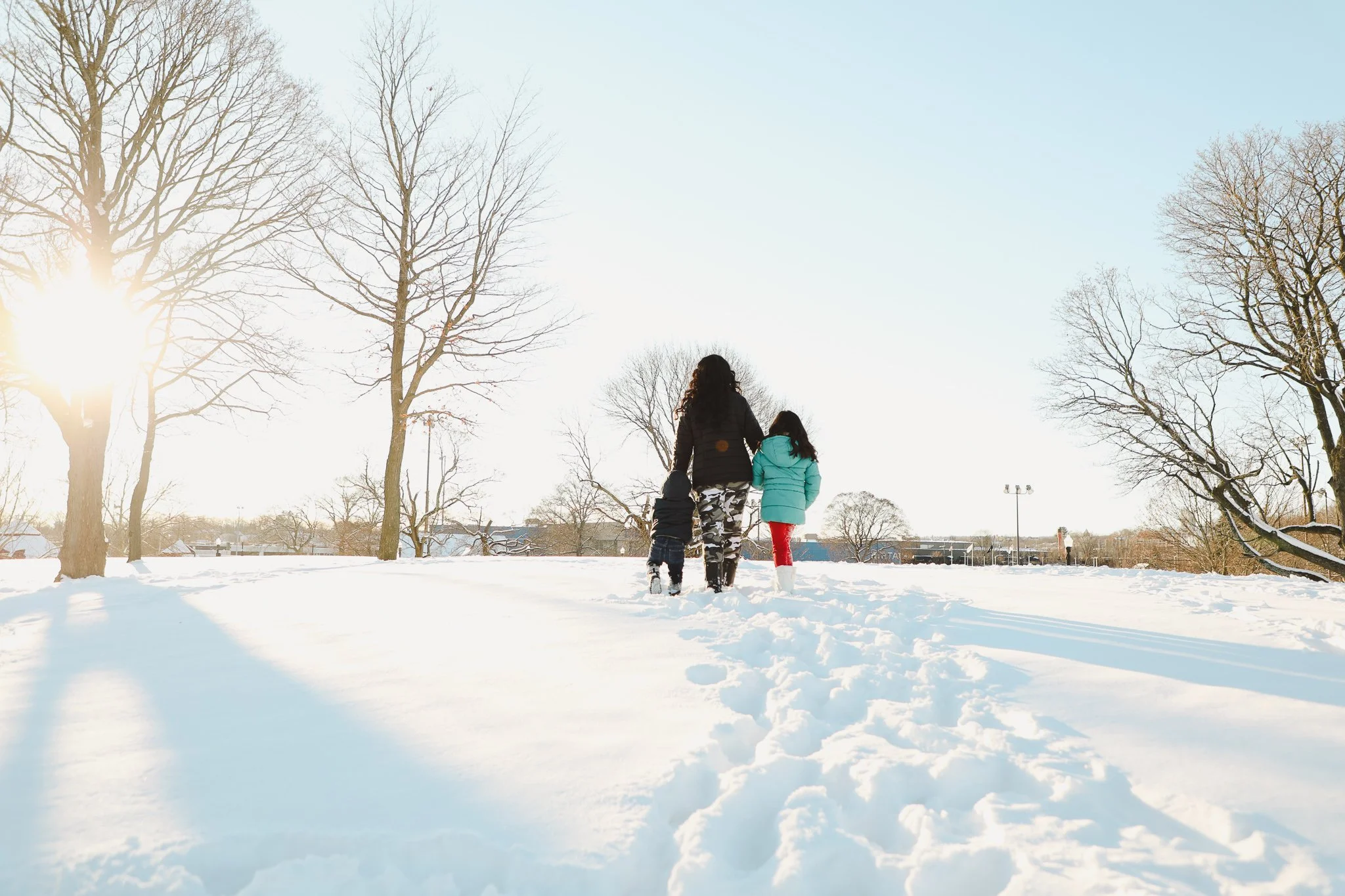 Family walking through snow-covered park on a sunny day, with leafless trees and a clear sky.