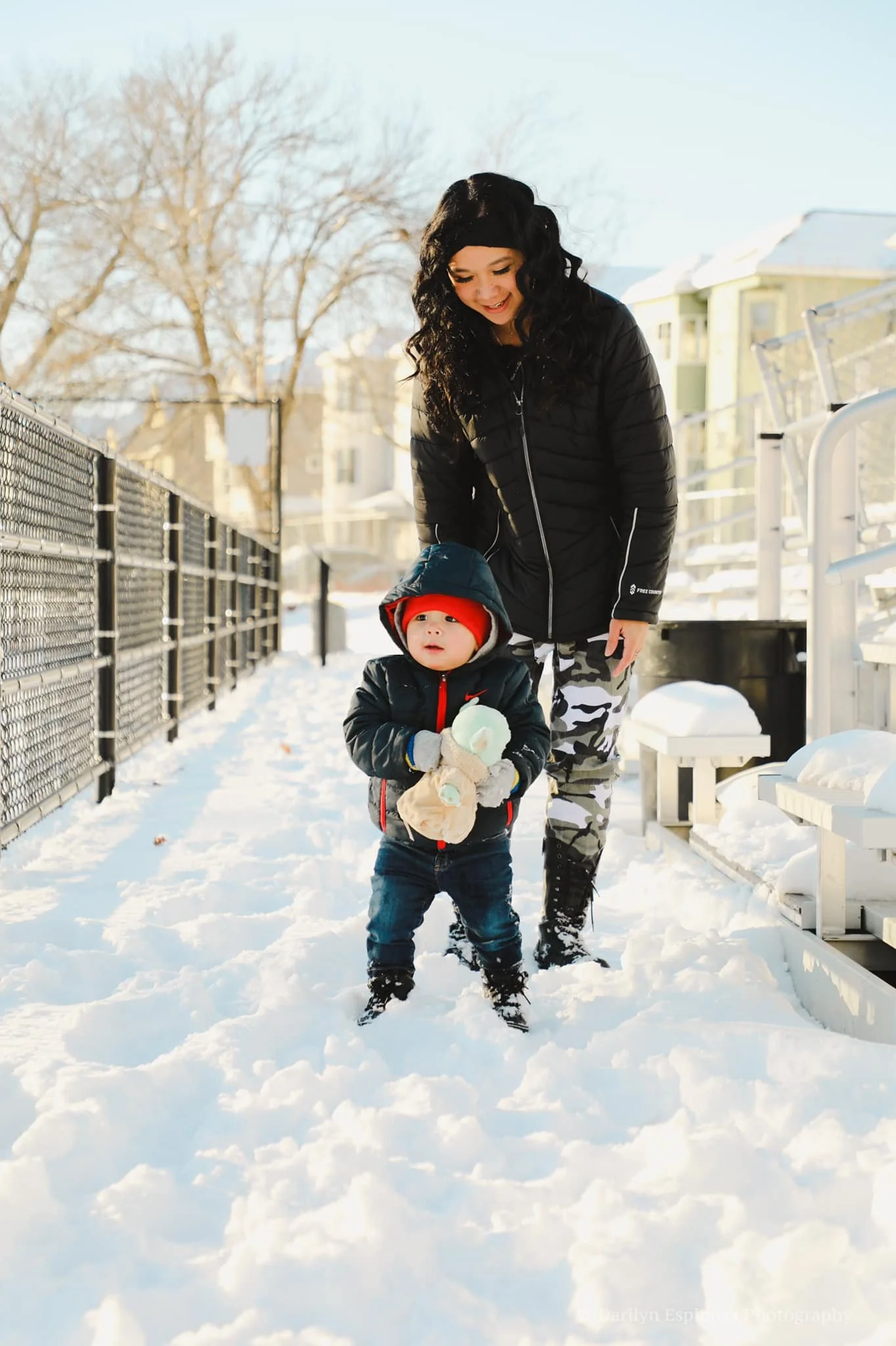 A woman and a young child in winter clothing walking through snow on a sunny day, with houses and trees in the background.