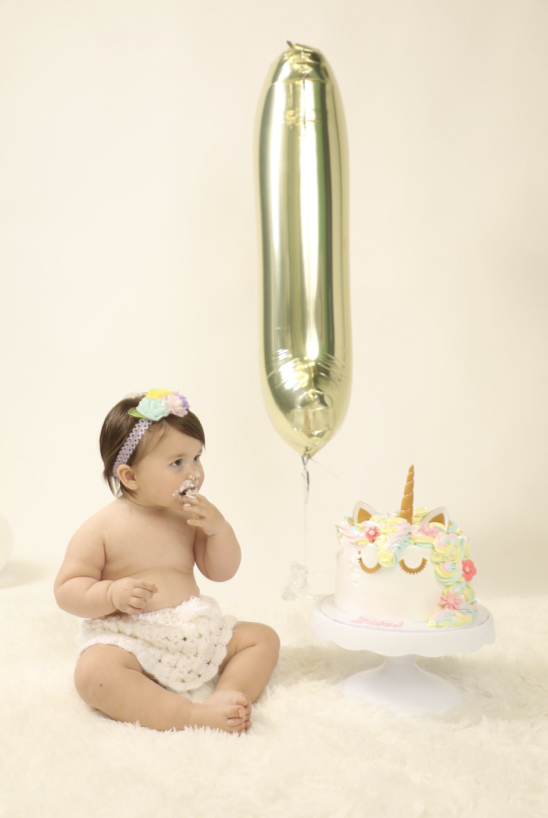 A baby girl with a flower headband sitting on a soft white surface, partially covered in a crocheted diaper, celebrating a first birthday with a unicorn cake and a large gold number one balloon in the background.
