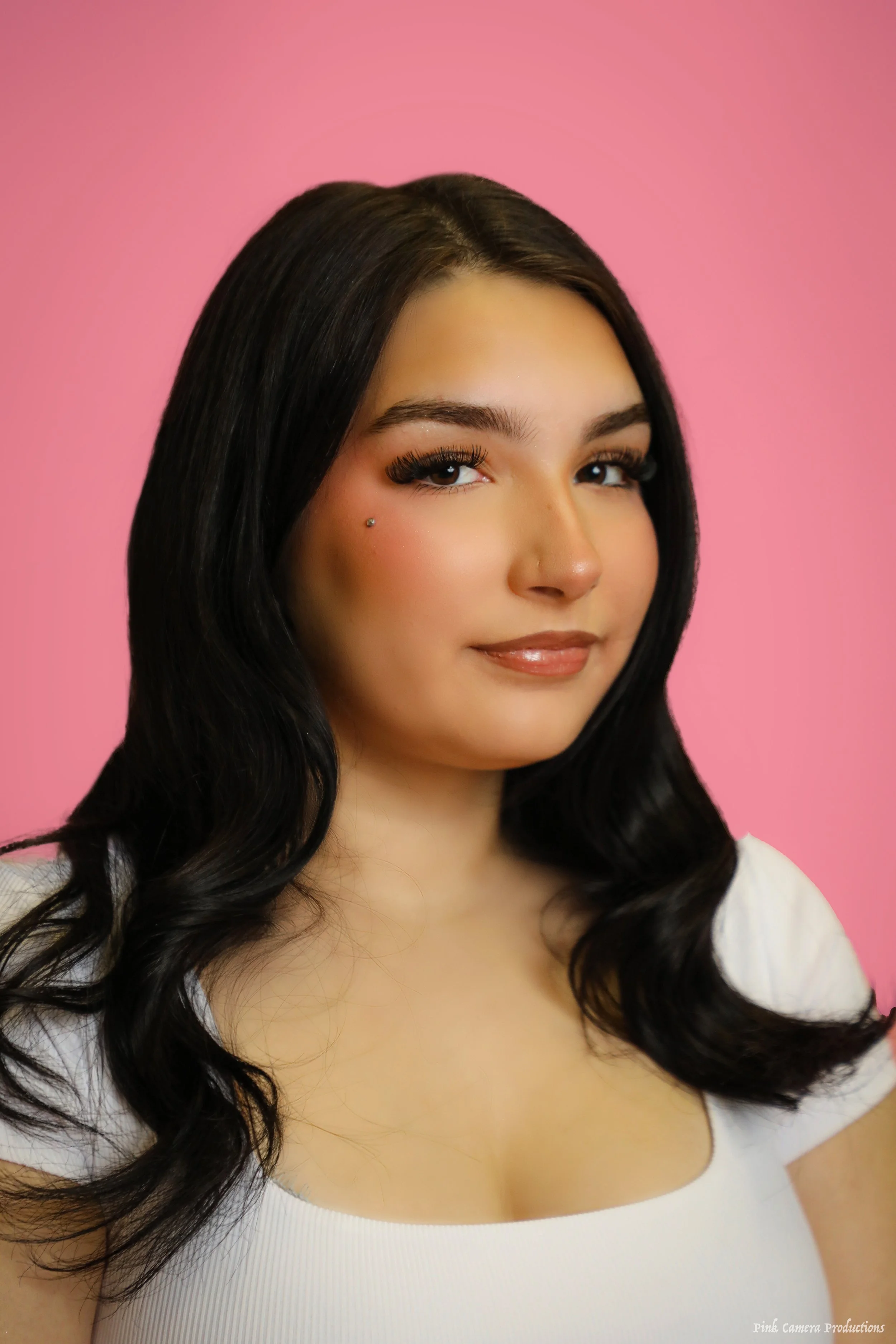 A young woman with long black hair, makeup, and a dermal piercing on her cheek, wearing a white top against a pink background.