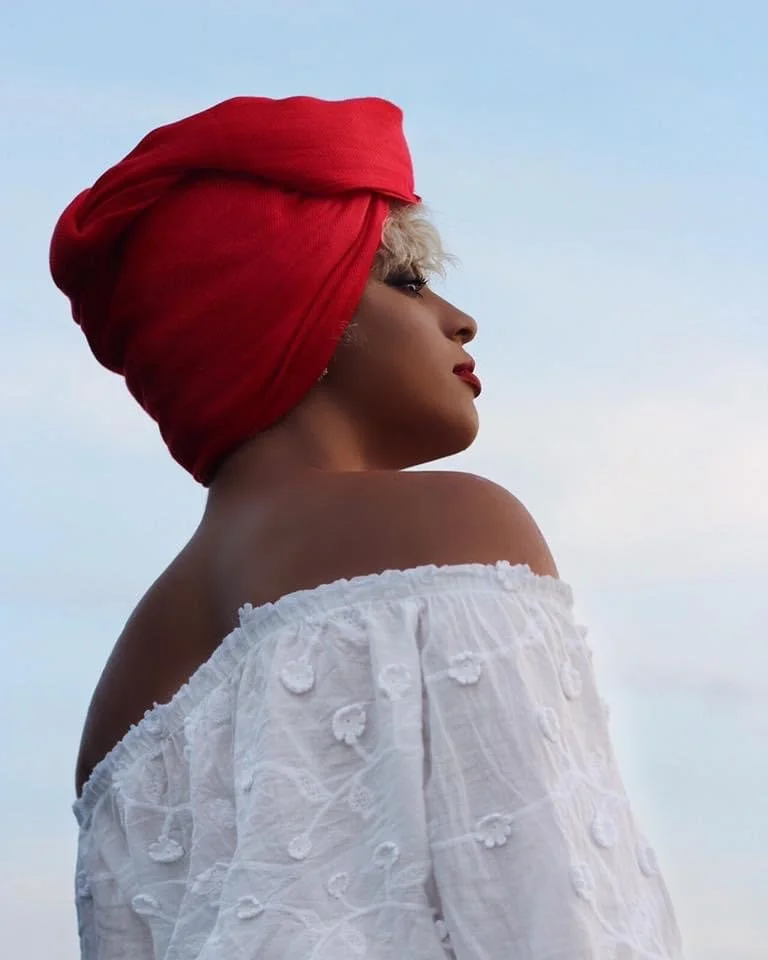 Profile of a woman with short blonde hair, wearing a large red headwrap and a white off-the-shoulder embroidered top against a sky background.