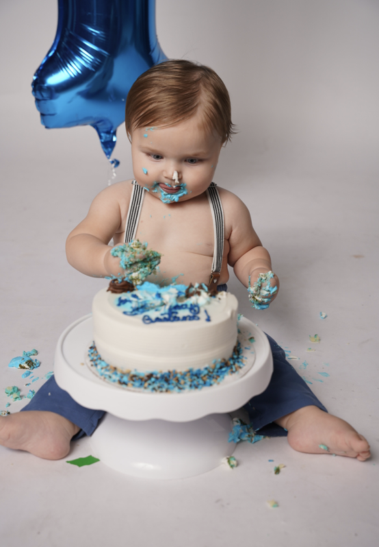 A young child celebrating a birthday, sitting shirtless with suspenders, eating a cake with blue and white frosting, surrounded by pieces of cake on the floor. There is a blue balloon in the background.