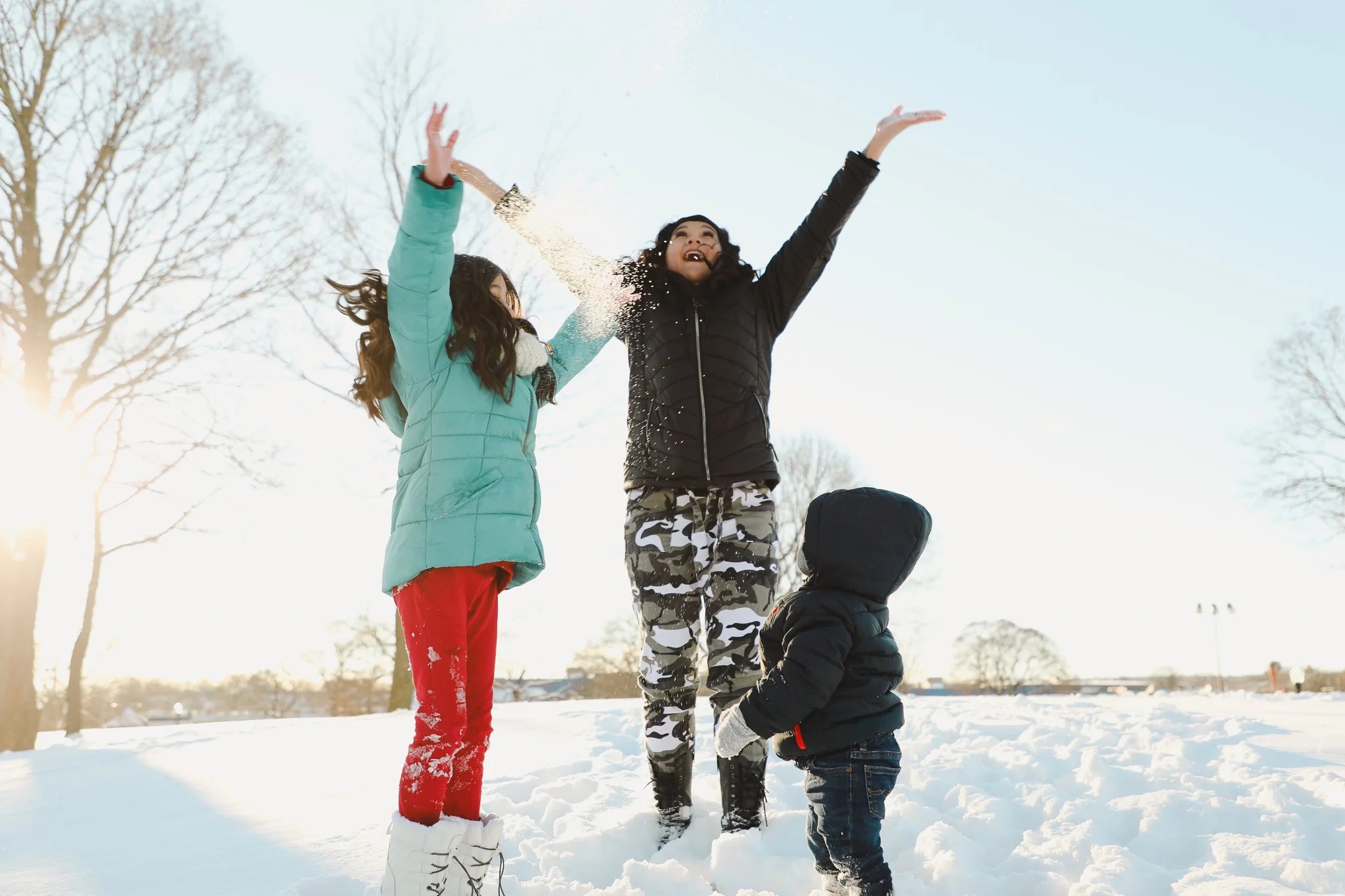 Three people playing in the snow outdoors during daytime, with two children and an adult woman, all dressed warmly in winter clothing.