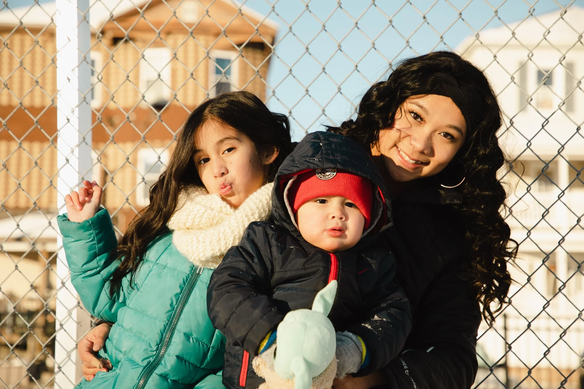 A woman with long, curly black hair is leaning in with two children against a chain-link fence on a sunny day. The girl on the left is making a playful face with pouted lips, wearing a teal winter coat and a cream-colored scarf. The boy in the middle