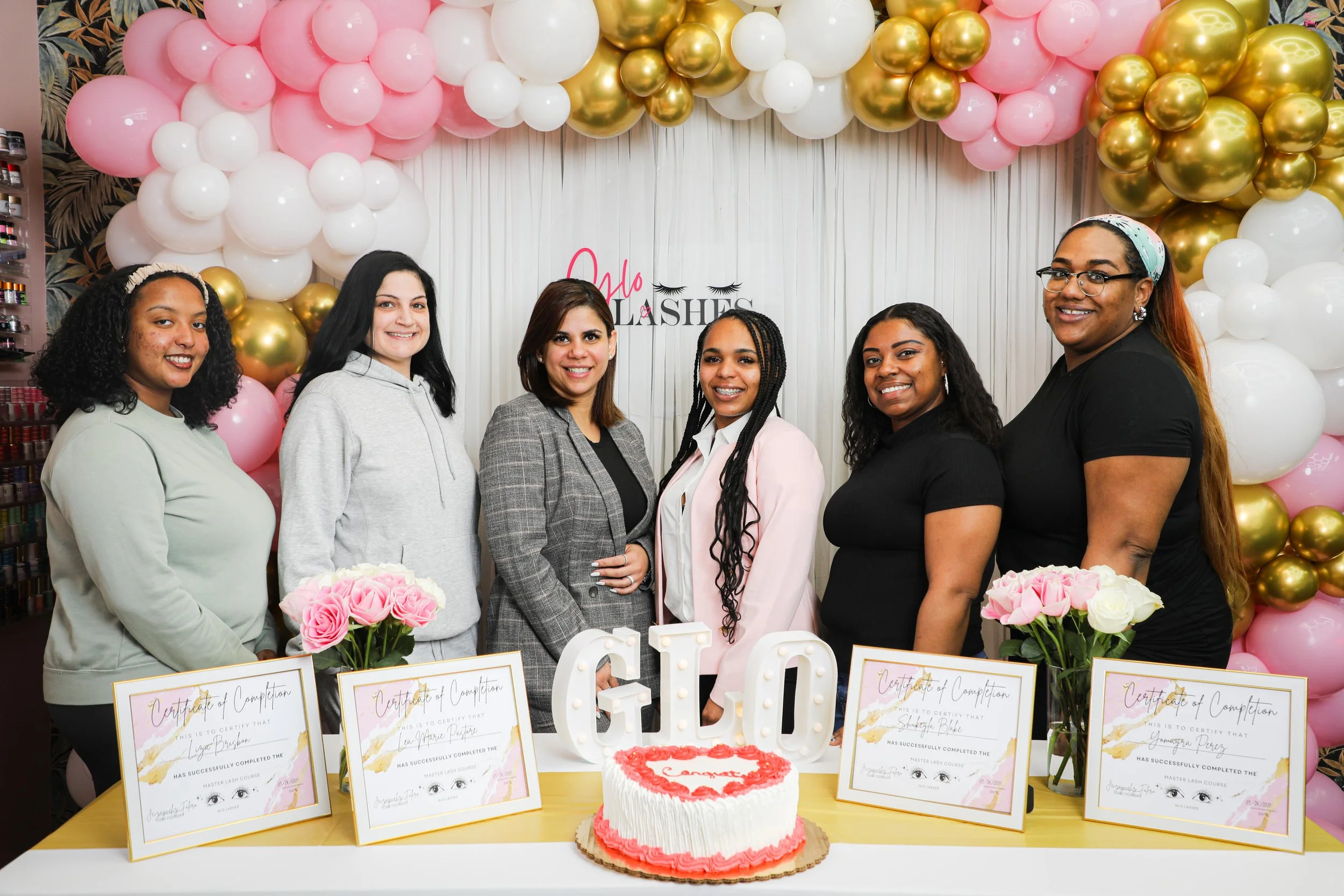 Six women at a celebration with pink, white, and gold balloons, a cake, certificates, and pink roses. The cake has bulbs forming the letters 'GLO' and a red heart on top. The women are smiling and standing behind a table with certificates and flowers