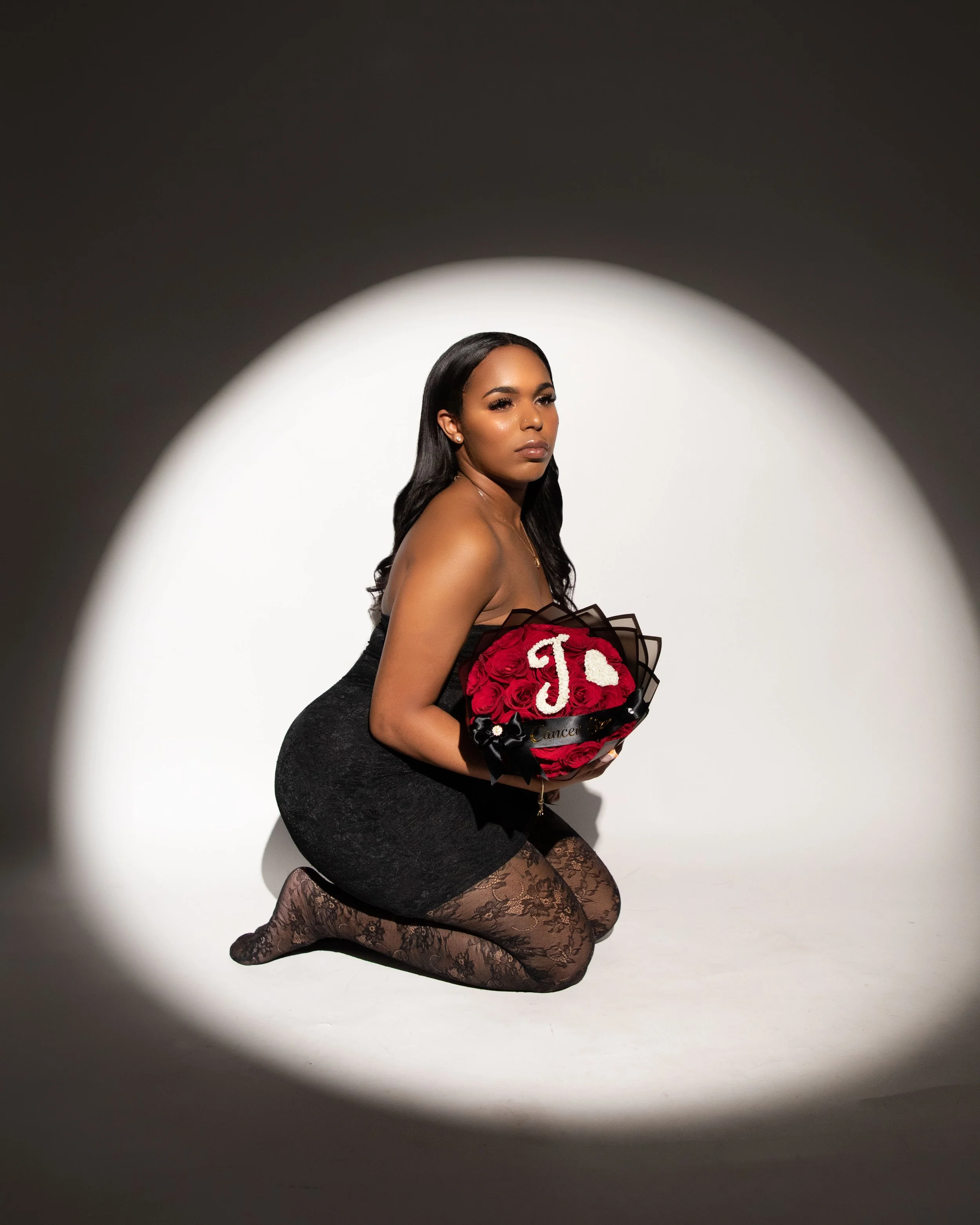 A woman kneeling on the floor holding a bouquet of red roses with a white heart and the letter 'J' on it, in a spotlight against a dark background.
