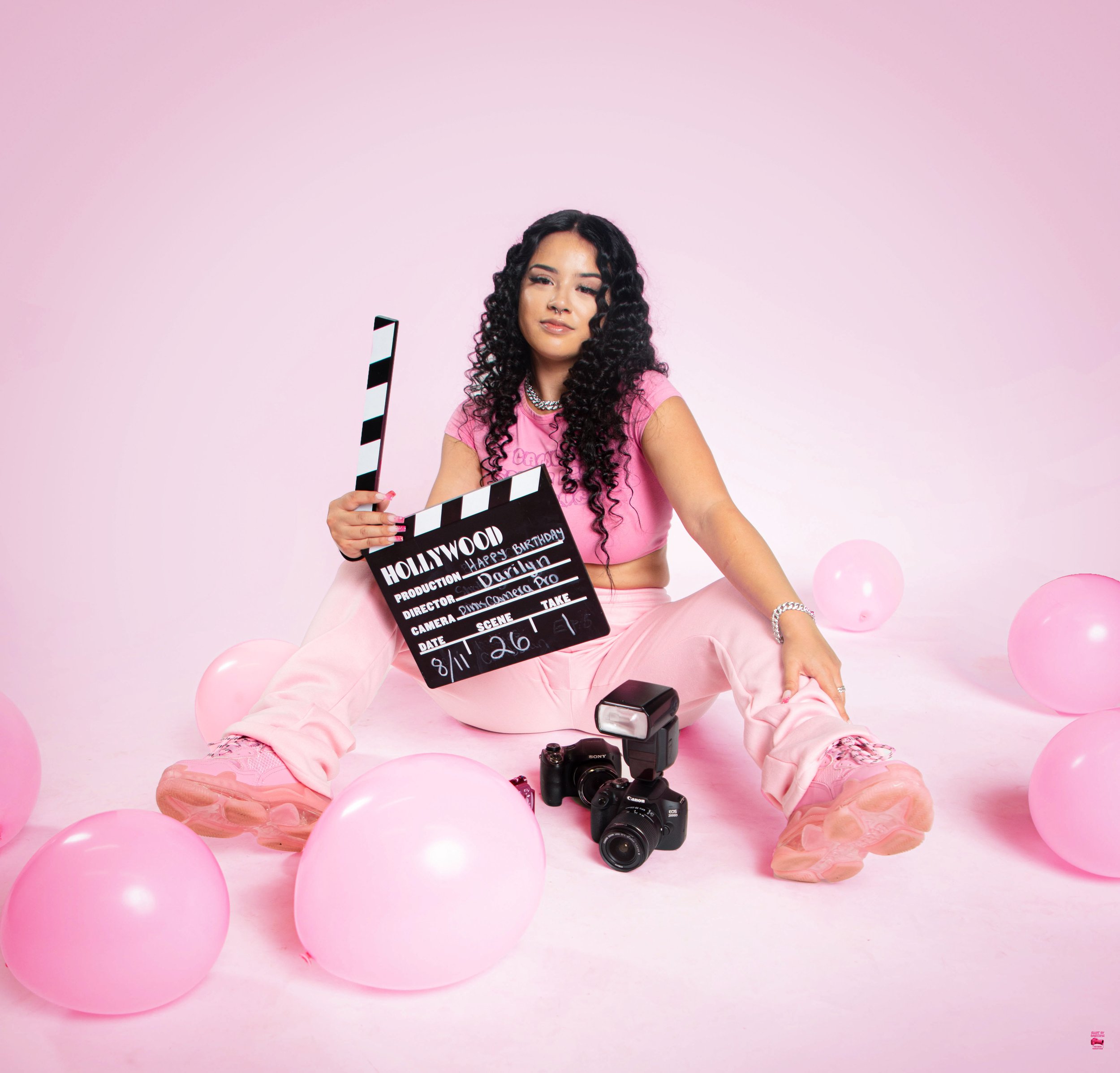 Young woman in pink outfit sitting on pink background surrounded by pink balloons, holding a clapperboard labeled Hollywood, with cameras and a flash nearby.