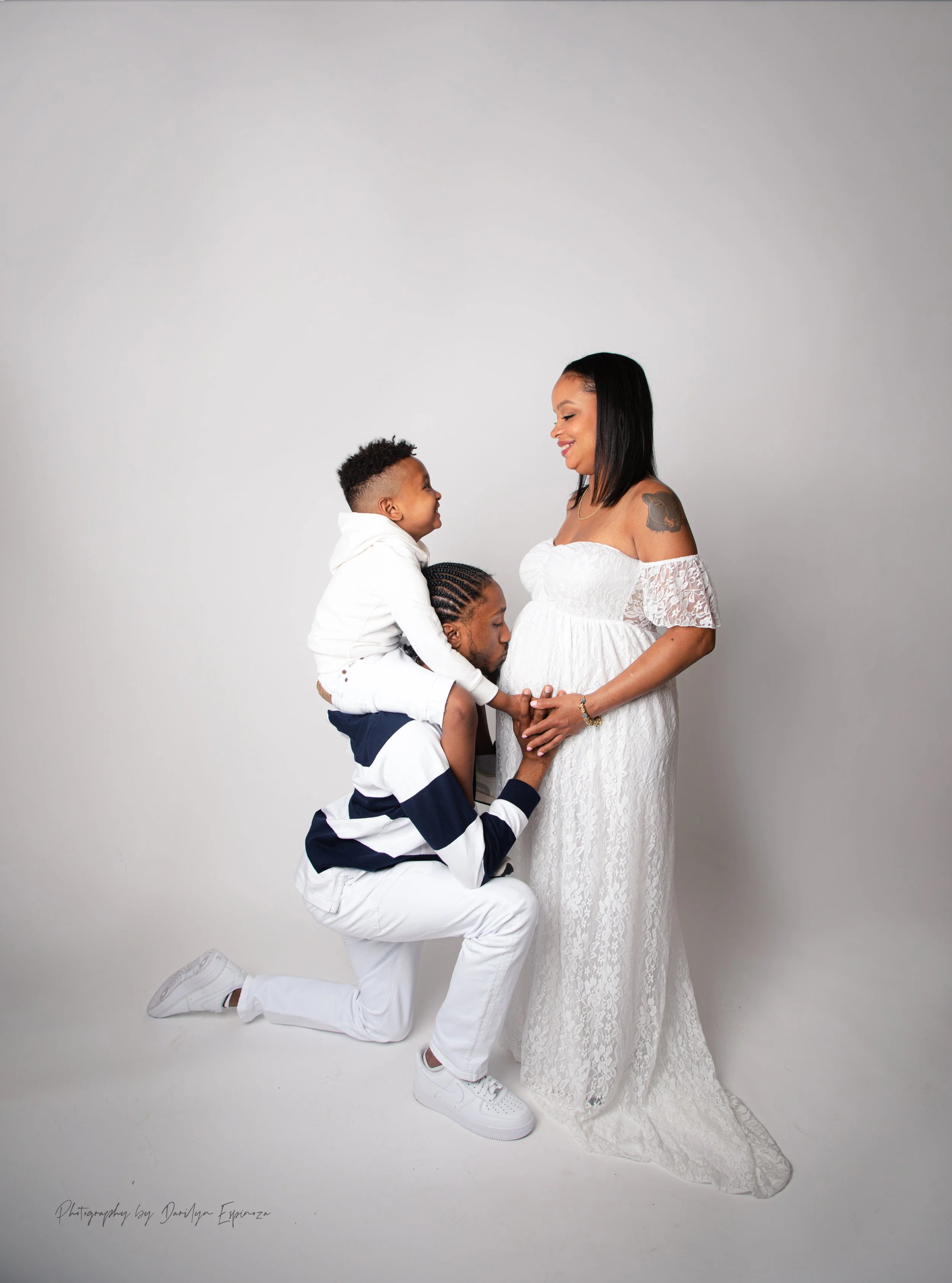 A pregnant woman in a white lace dress smiling at a man kneeling, holding her belly, with two children, one sitting on his shoulders and the other standing, all facing each other in a studio setting.