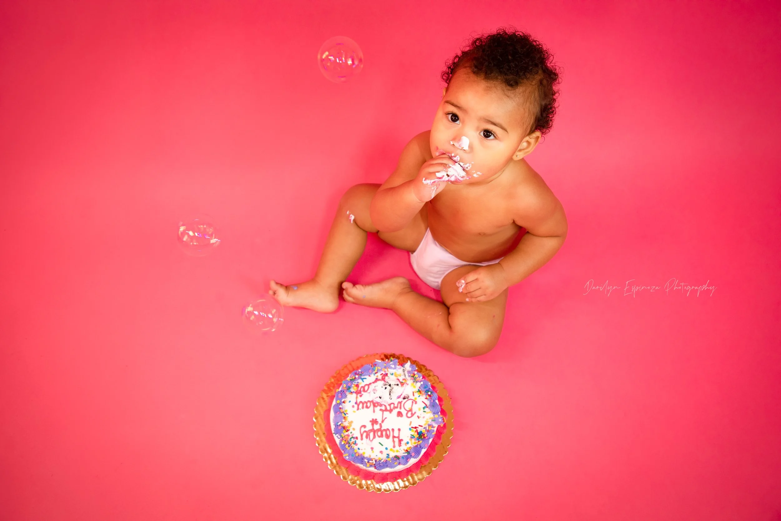 A baby sitting on a pink background with a birthday cake, blowing bubbles, and smeared cake on face and hands, celebrating a first birthday.