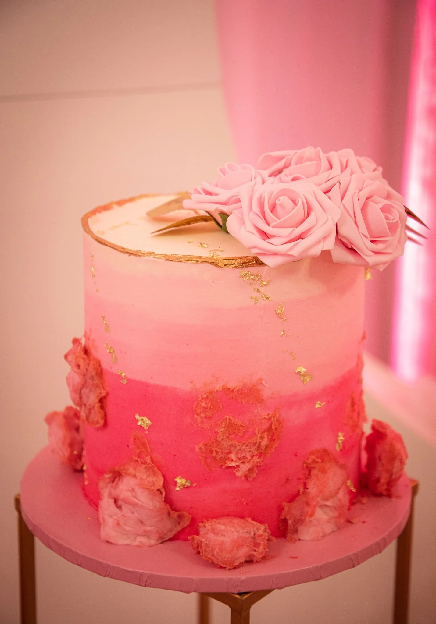 Pink and white ombré cake decorated with pink roses and gold accents, placed on a pink cake stand.