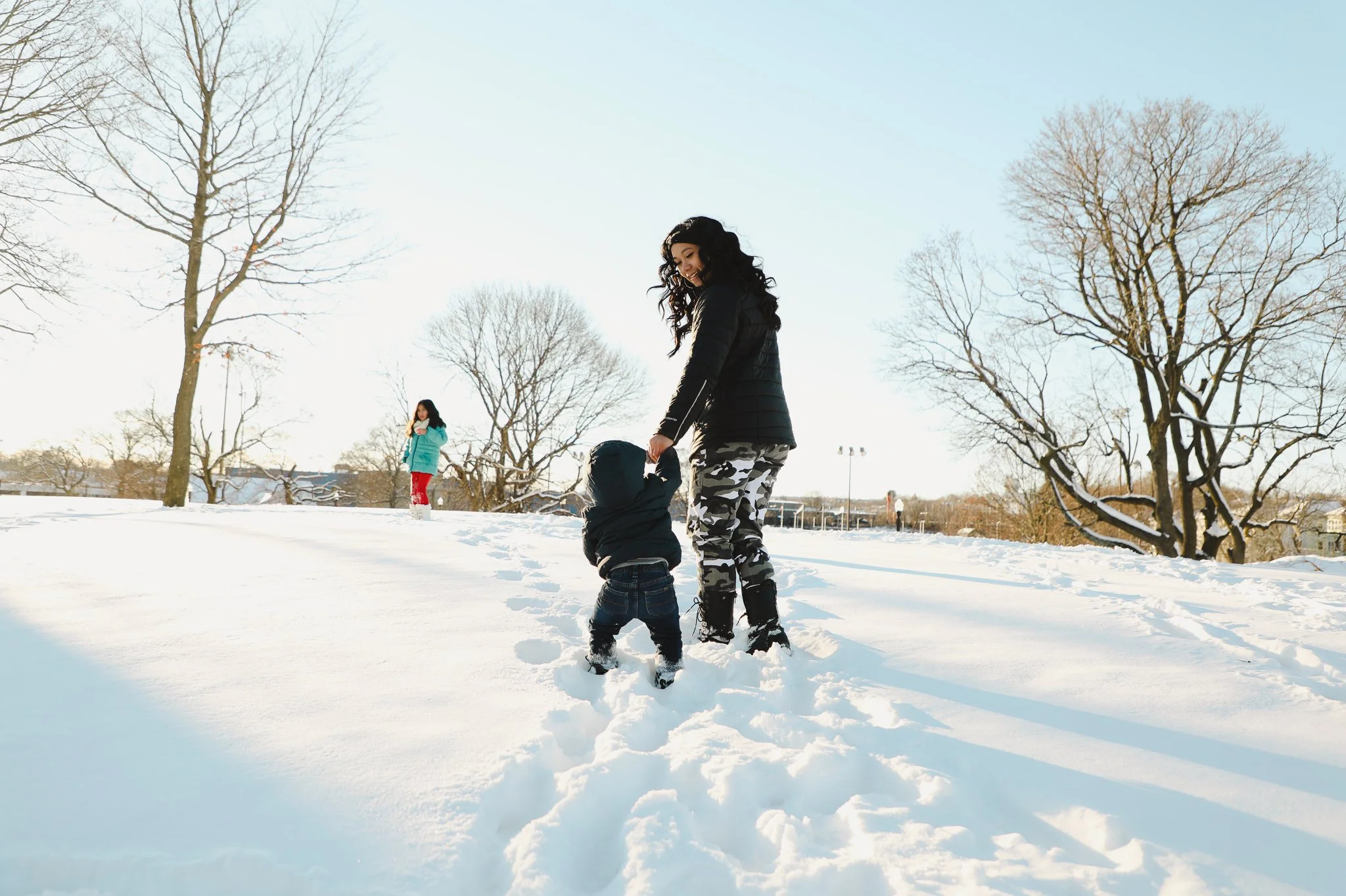 A woman holding hands with a young child in a snowy park with leafless trees, another child in the background, and a clear blue sky.