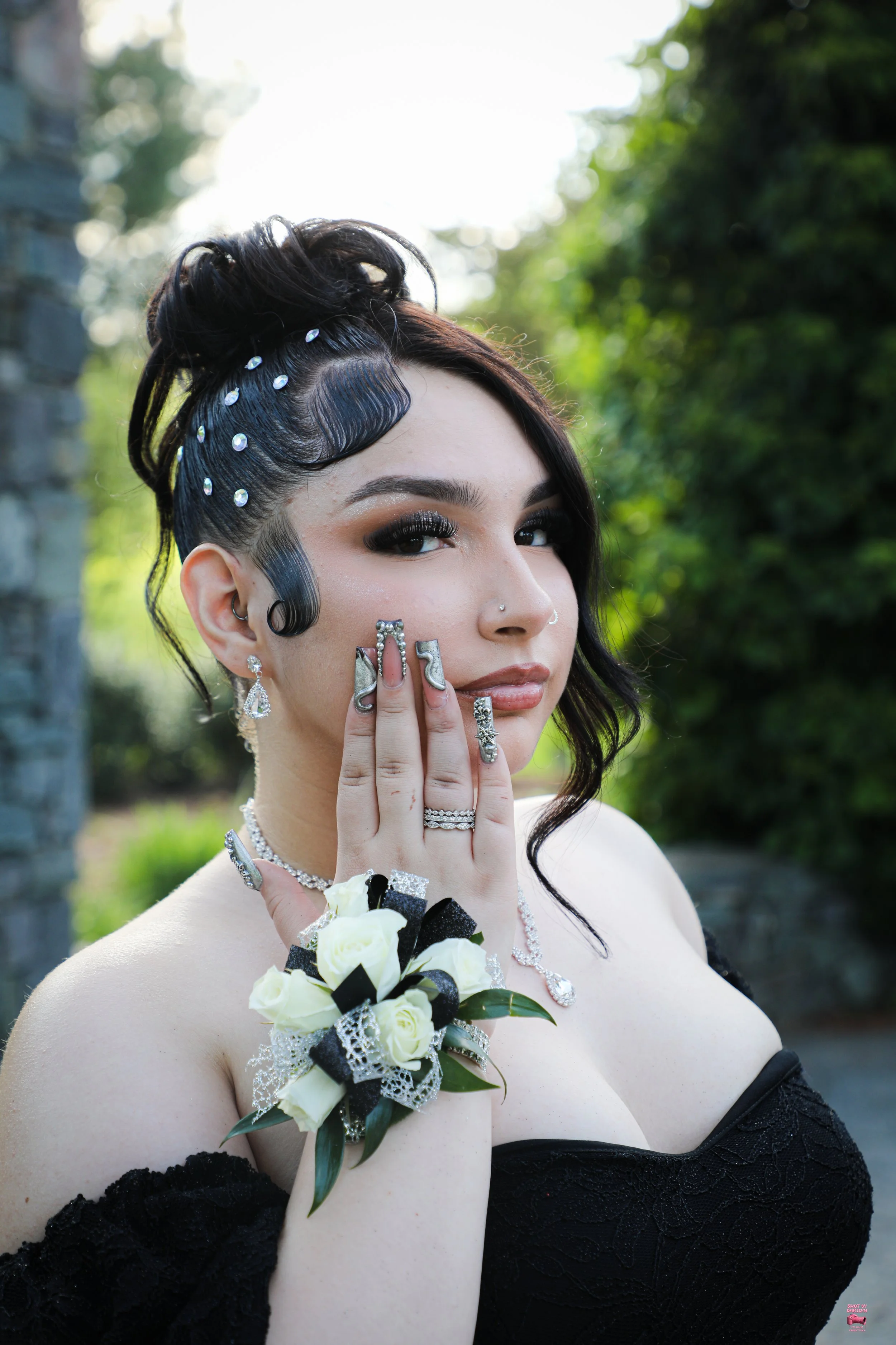 A woman with dark, styled hair and multiple rhinestone hair accessories, wearing a black off-shoulder dress, multiple rings, earrings, and a white flower corsage. She has elaborate makeup with dark eye shadow and nude lipstick, standing outdoors with