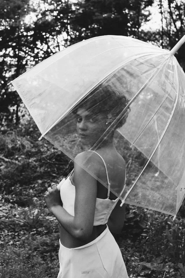 A young woman with short hair, wearing a sleeveless top and skirt, holds a clear umbrella and stands outdoors among trees and foliage.