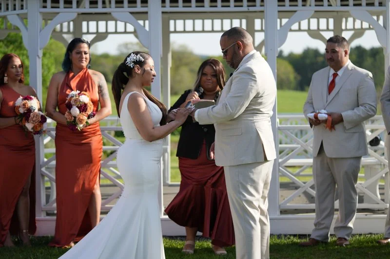 Bride and groom exchanging vows at outdoor wedding ceremony, with bridesmaids and groomsmen in the background.