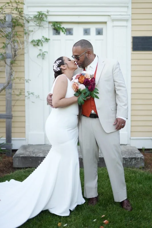 A newlywed couple sharing a kiss outdoors. The bride is in a white wedding dress with a train and holding a bouquet of flowers. The groom is in a light-colored suit with a red vest and dark shoes. They are standing on grass in front of a white buildi