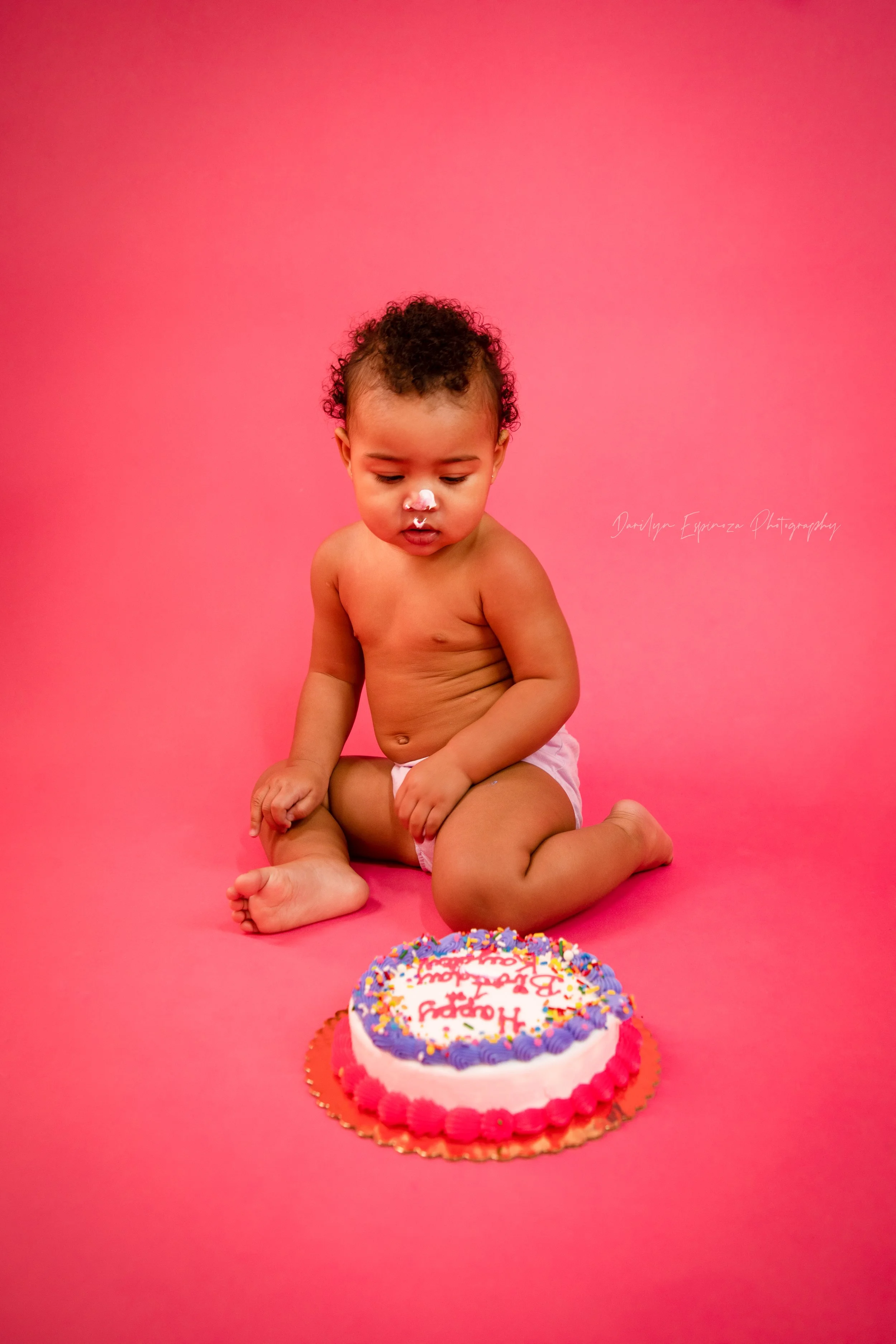 A young child with curly hair, wearing only a diaper, sitting on a pink background, celebrating their birthday with a cake in front of them.