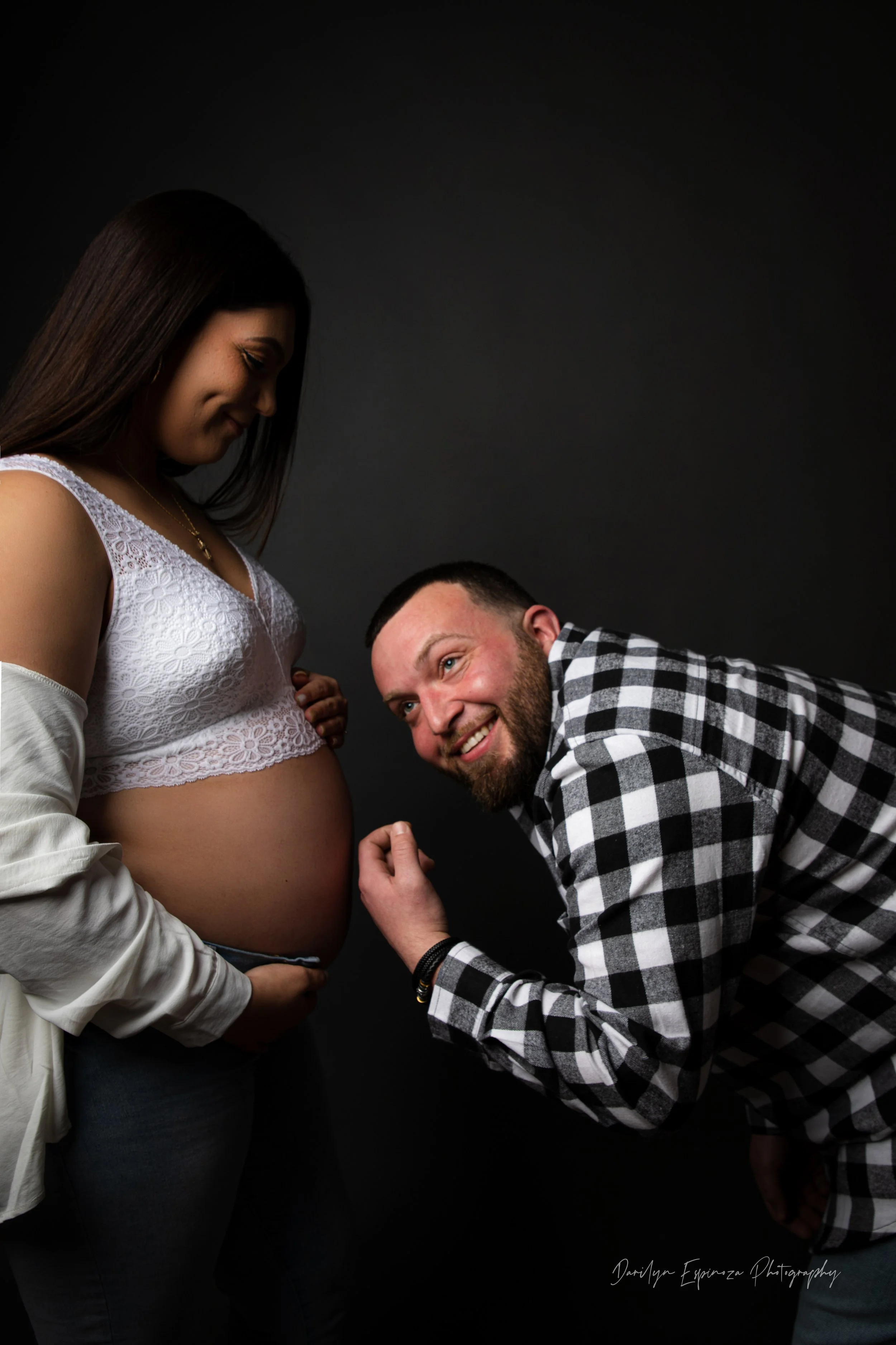 A man and a pregnant woman posing for a maternity photo against a dark background. The man is smiling and leaning toward the woman's belly, which she is gently holding with one hand. She is looking down at her belly, wearing a white lace top and a be
